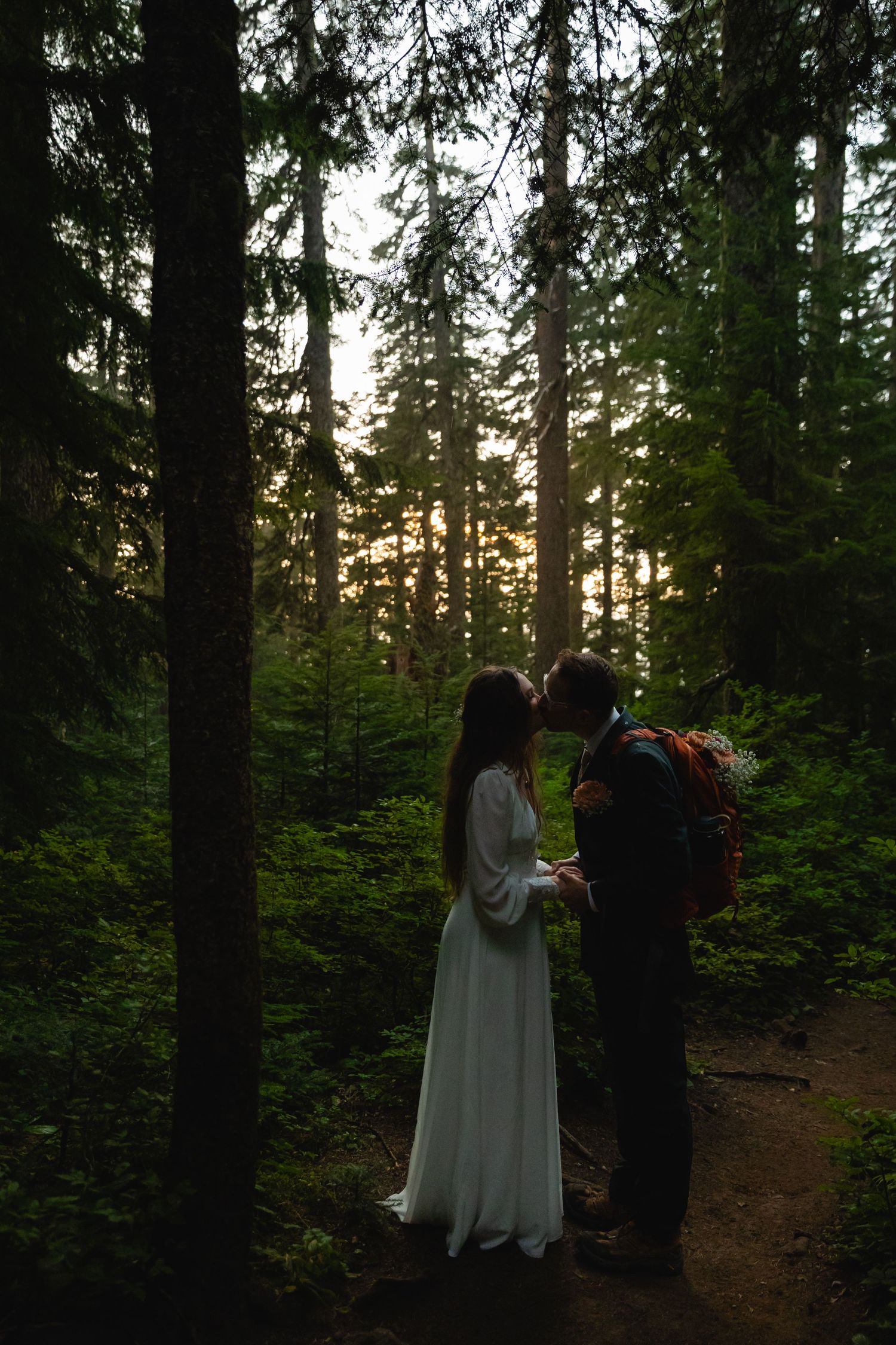 Wedding couple stands in silhouette against sunset light filtering through evergreen forest trees.