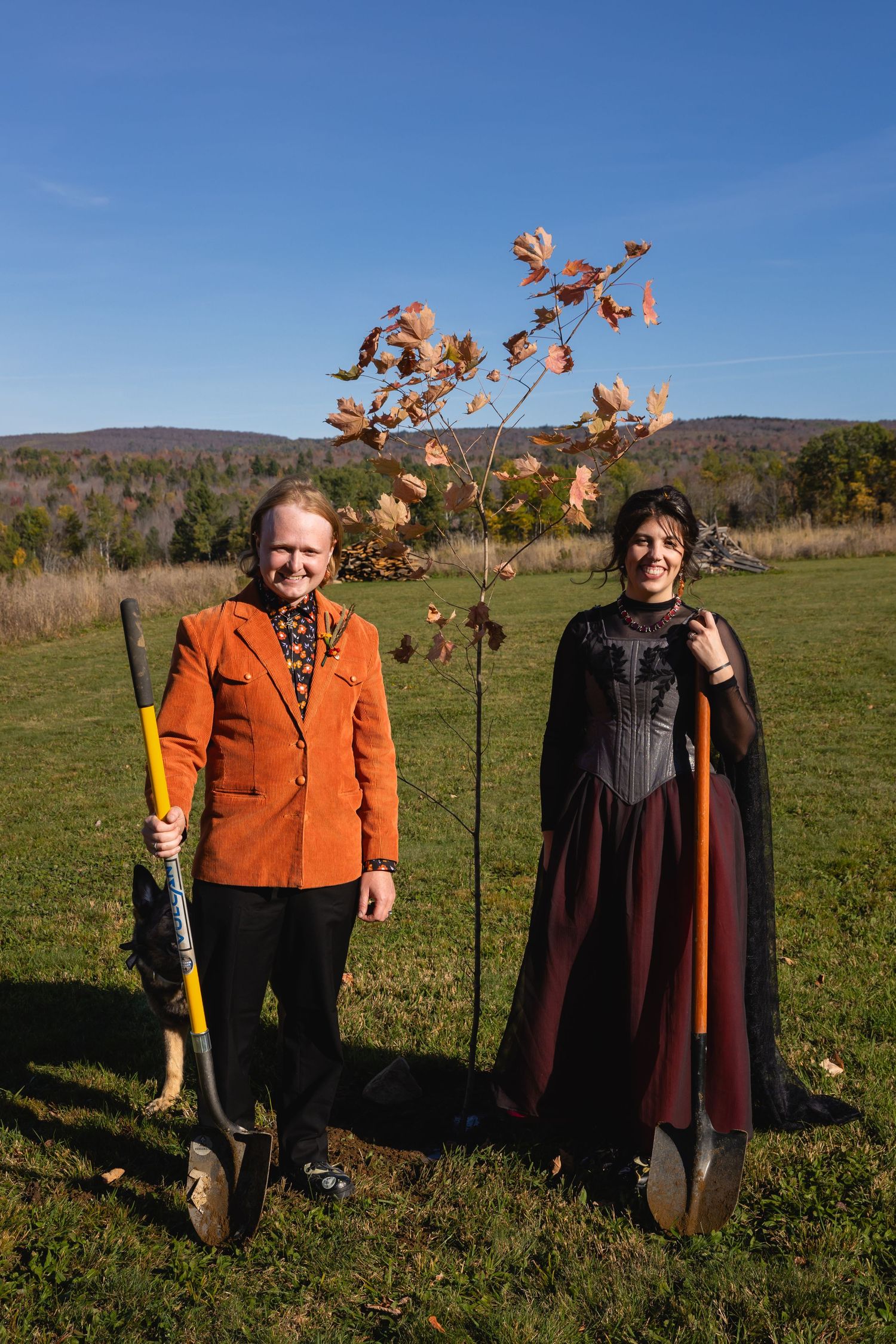 Two people stand beside newly planted trees in a rural field with mountains visible in the background.