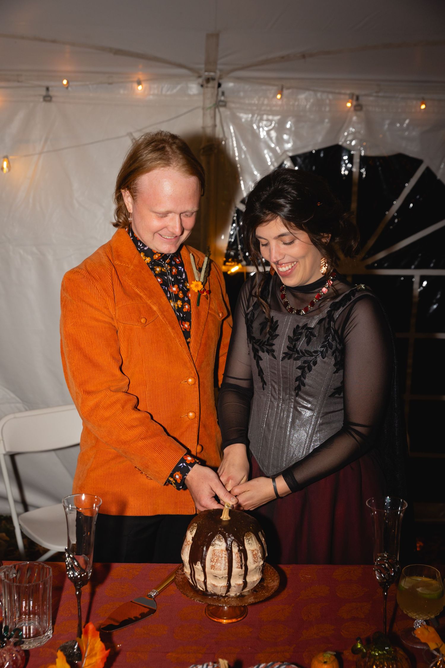 Two people in formal attire cut into a decorated cake at an indoor celebration with string lights overhead.