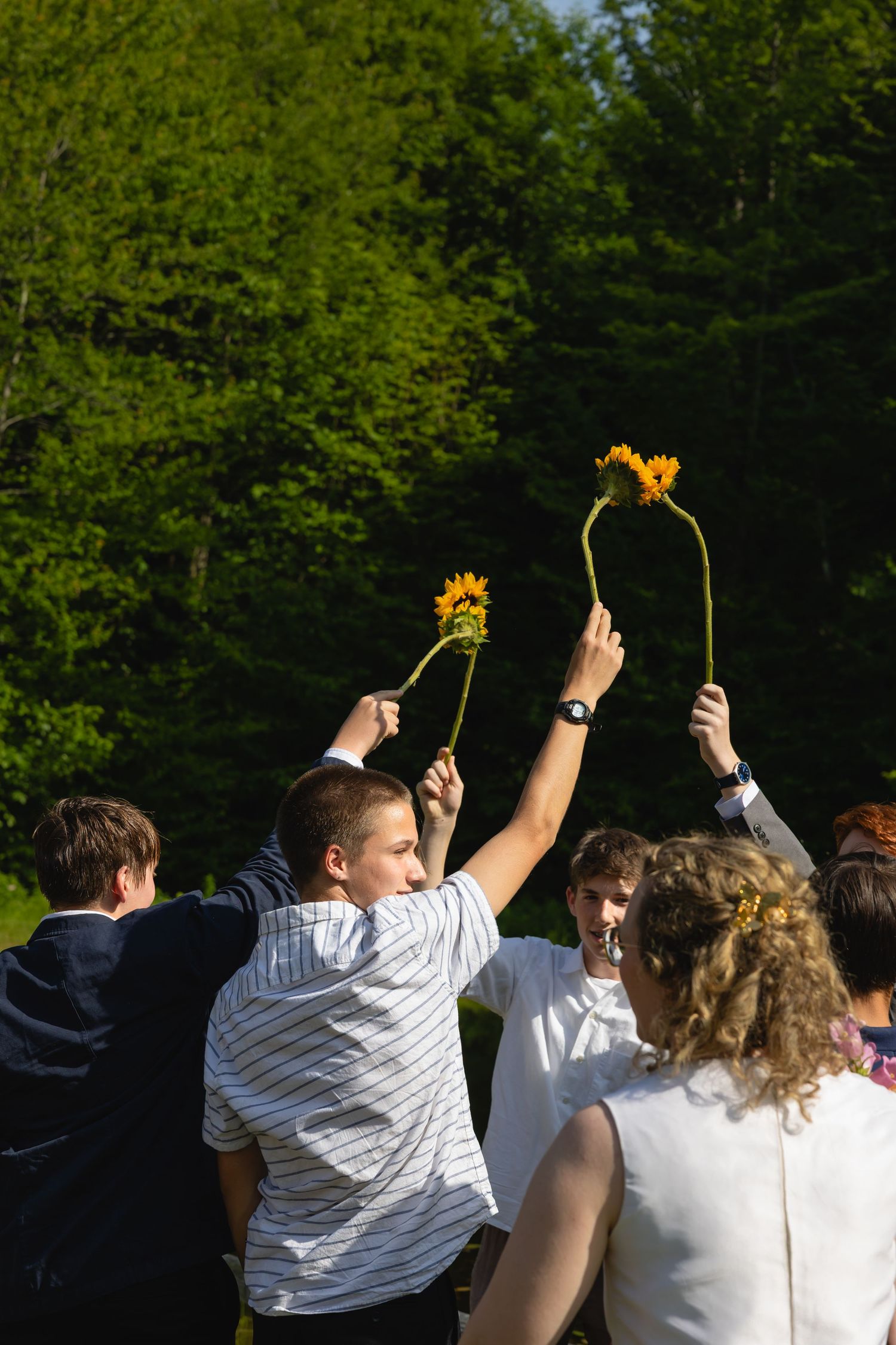 A group of people raise sparklers in celebration against a green forest backdrop during an outdoor gathering.