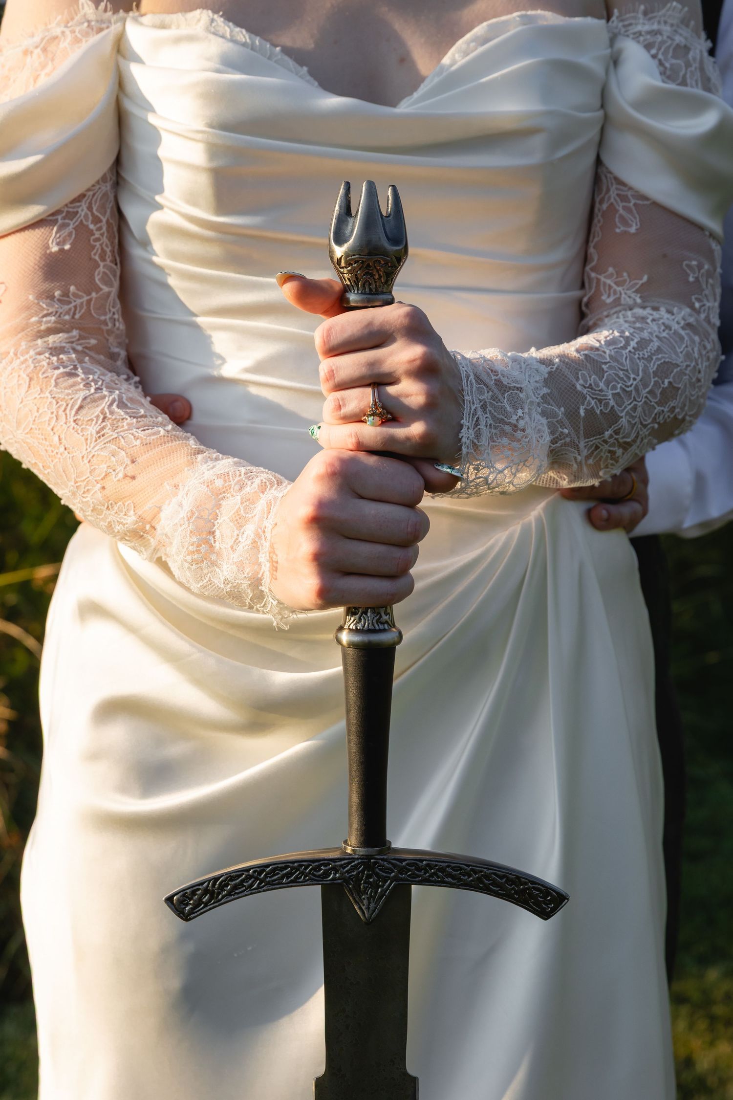 A person in a white wedding dress holds a decorative medieval sword against a sunlit outdoor background.