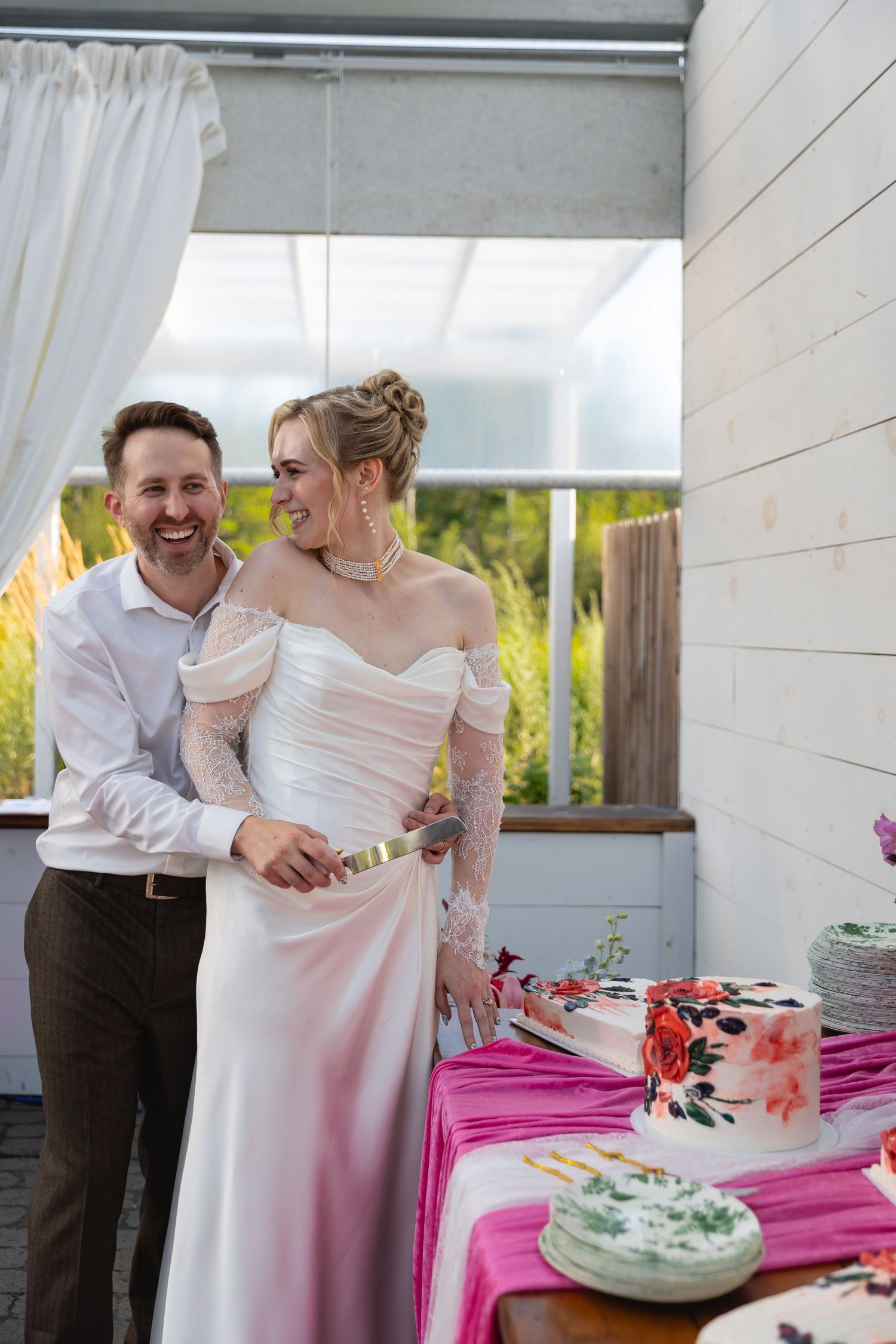 A couple shares a sweet moment while cutting their wedding cake together at an outdoor reception with pink decor.
