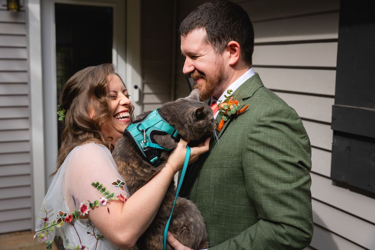 A couple shares a joyful moment while holding their gray cat wearing a teal bowtie during a special occasion.