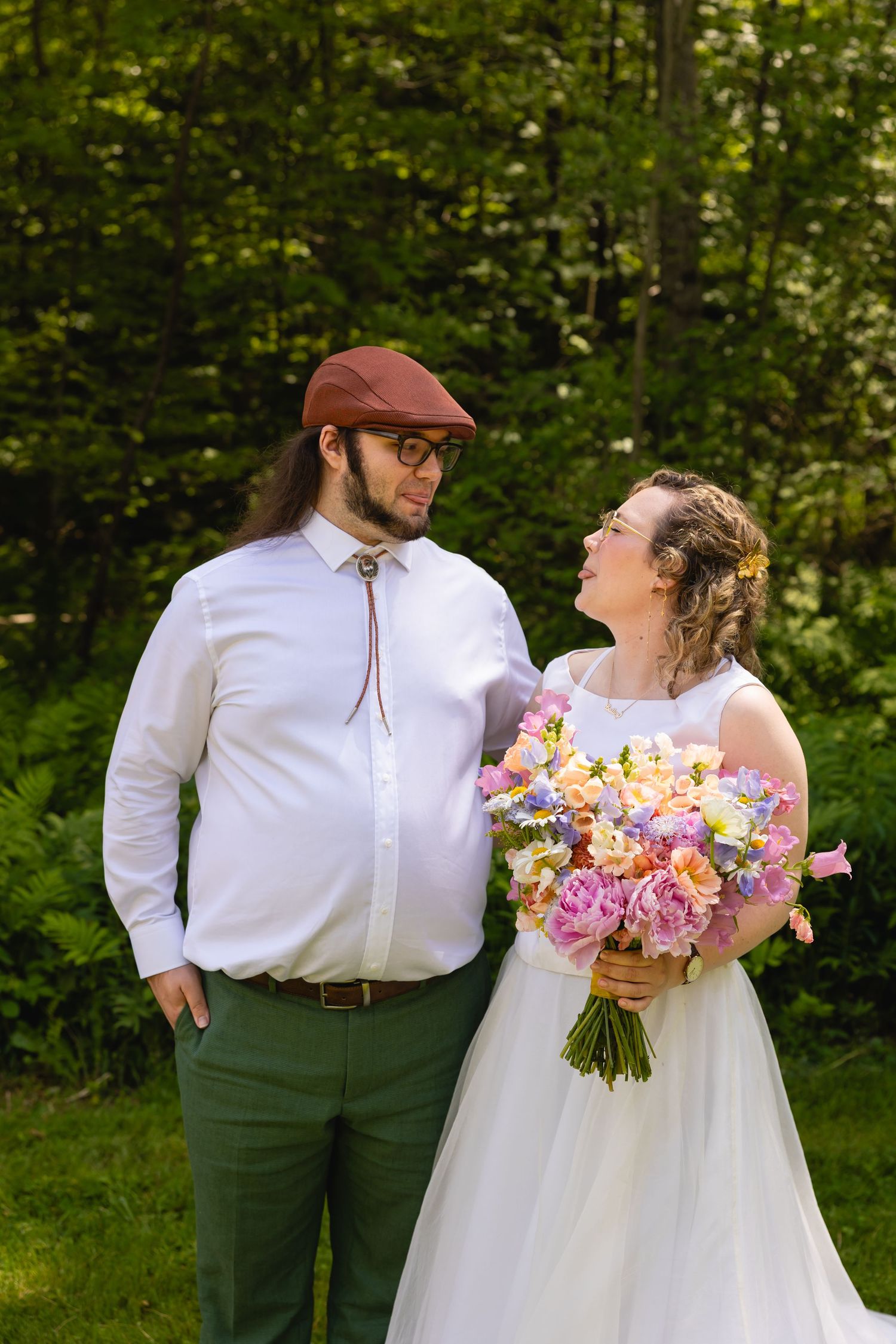 A joyful couple shares a tender moment outdoors in wedding attire with a colorful pink and peach floral bouquet.