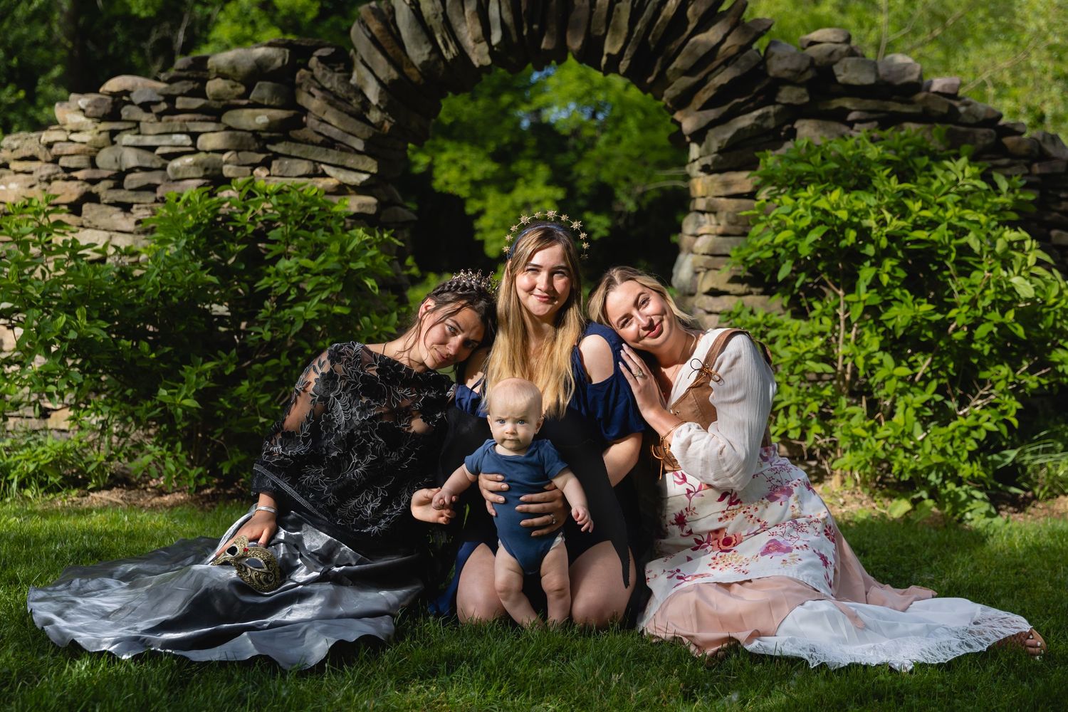 Three women and a baby posing together in garden with stone arch backdrop.