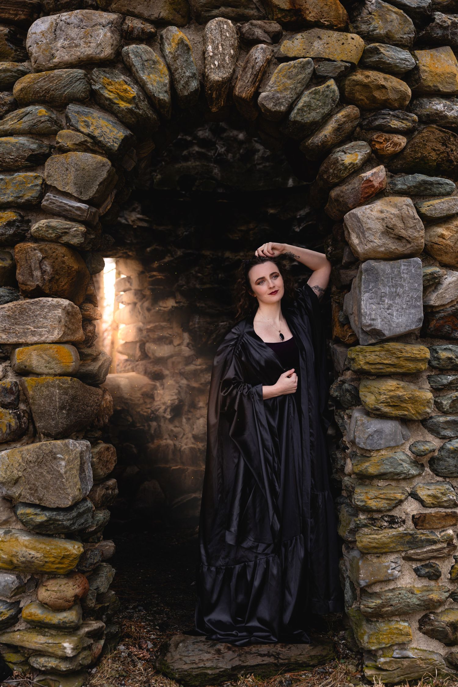 Woman in black gown standing in stone arch doorway with warm light glowing behind her.
