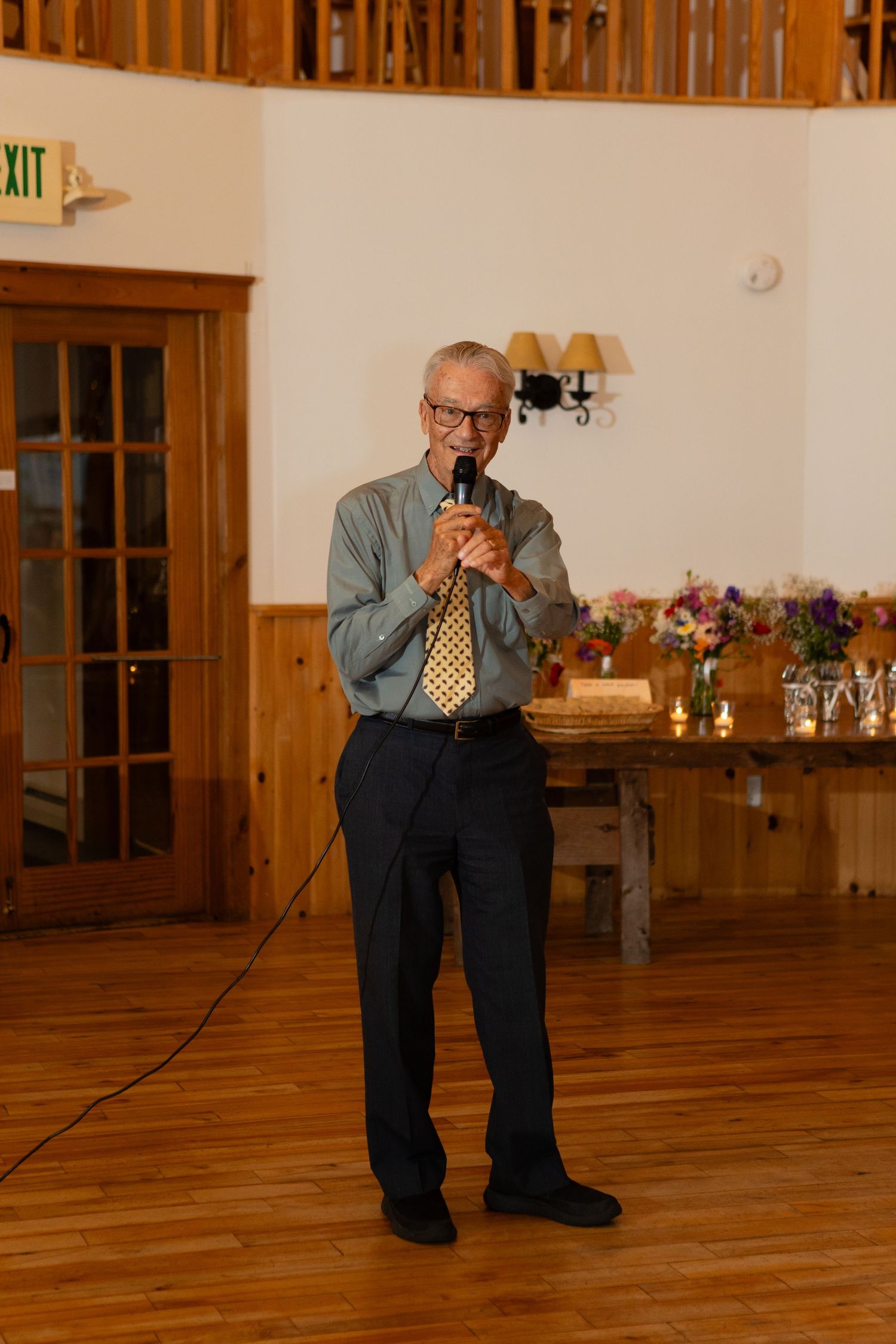 Speaker holding microphone during wedding reception in rustic wood-paneled venue.