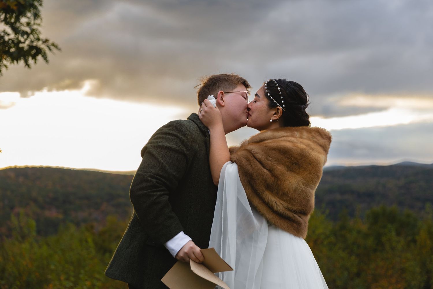 A romantic wedding kiss against a backdrop of mountain scenery at sunset with the bride wearing a fur wrap.
