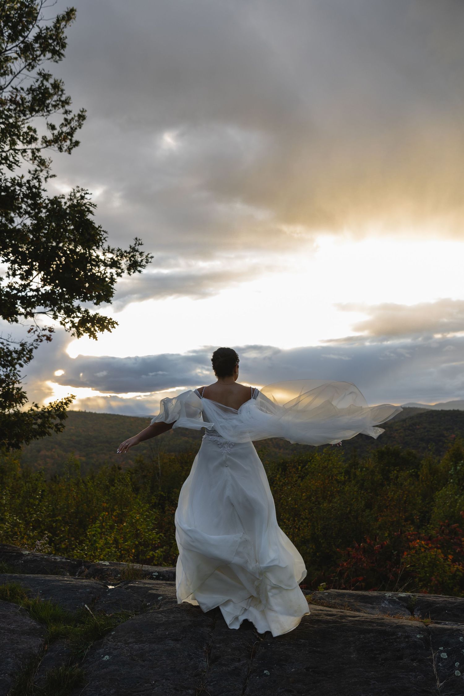 A silhouetted figure in a wedding dress stands with arms outstretched against a mountain sunset.
