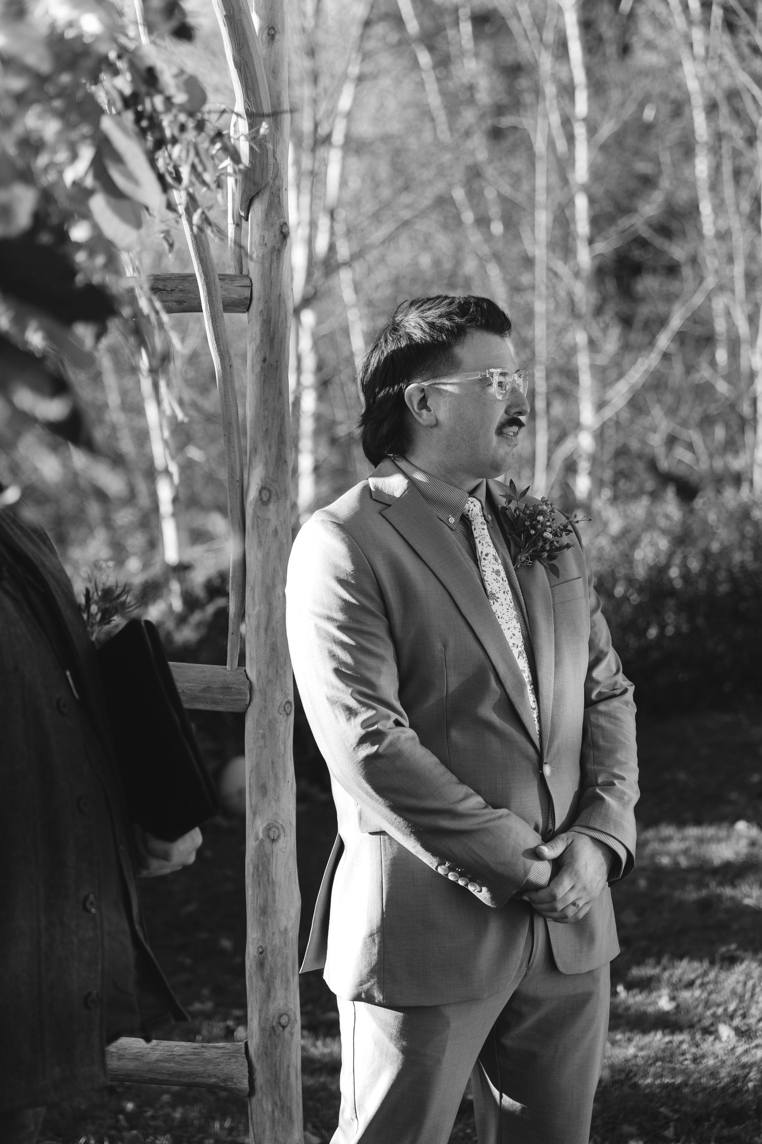 Wedding ceremony participant in tan suit standing outdoors among birch trees.