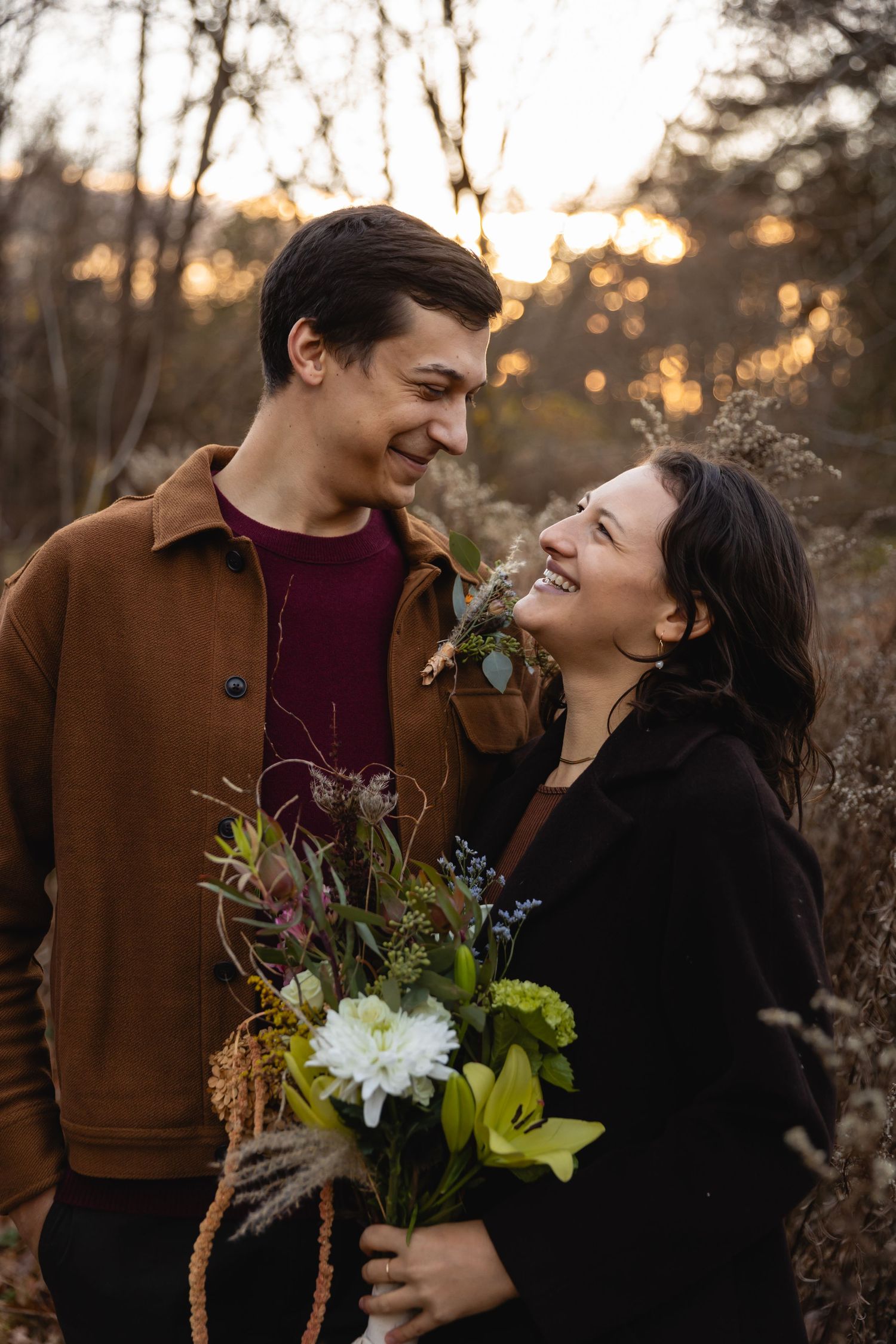 A couple shares a candid laugh during a sunset engagement photoshoot while holding a bouquet of wildflowers.