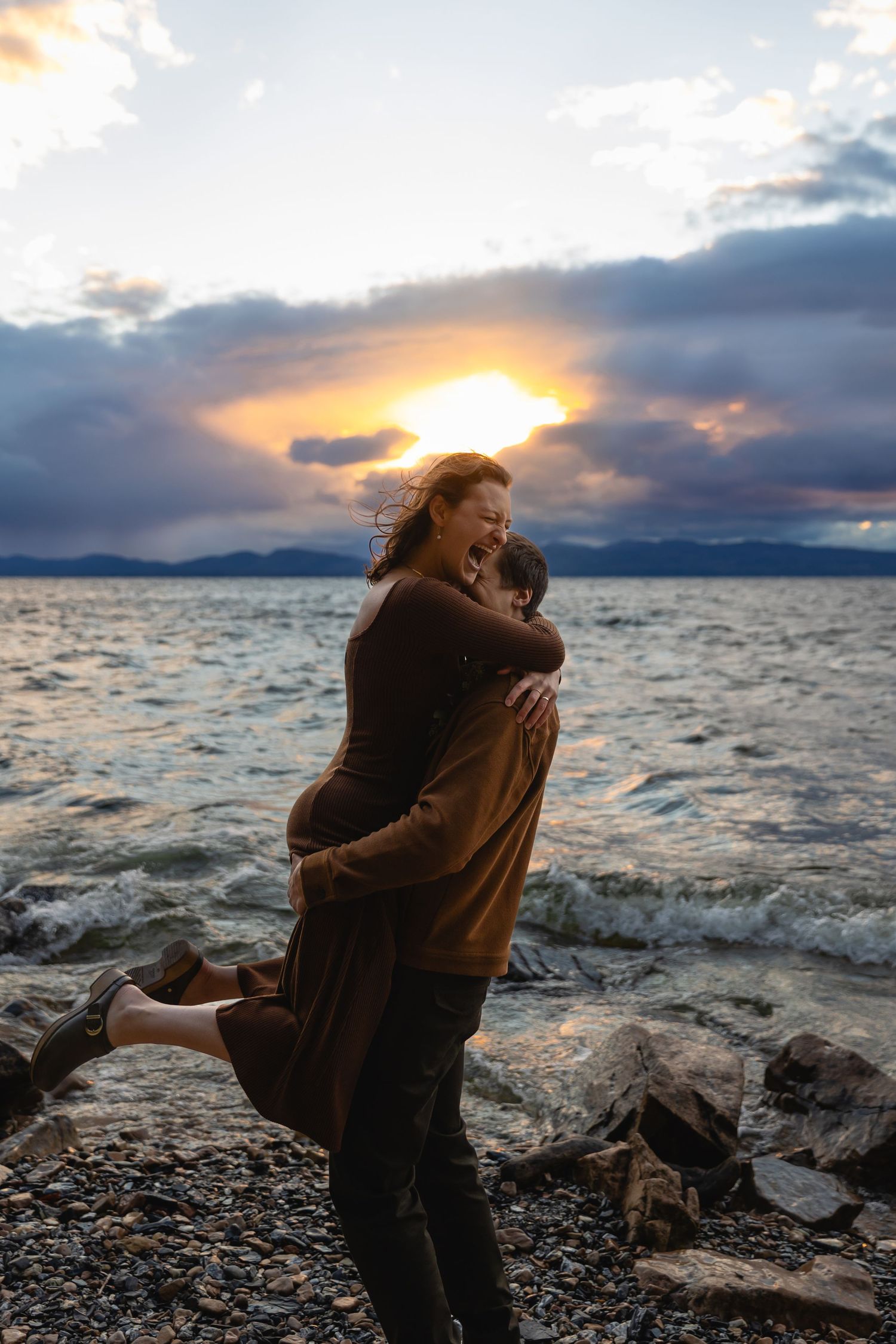 A couple embraces on a rocky beach shore during sunset with dramatic cloudy skies over the water.