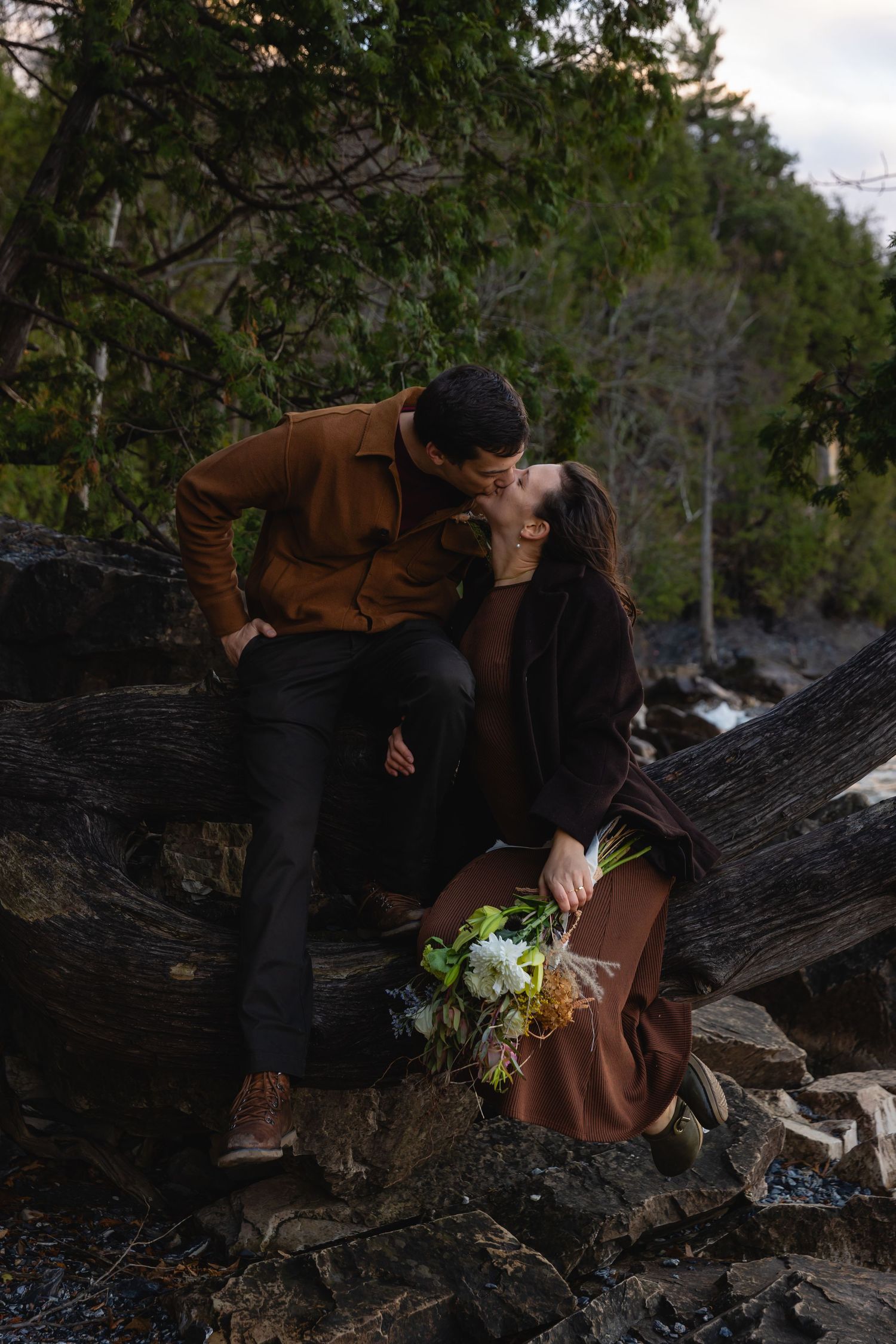 A romantic couple sits on driftwood at dusk, dressed in brown earth tones with a white floral bouquet against forest backdrop.