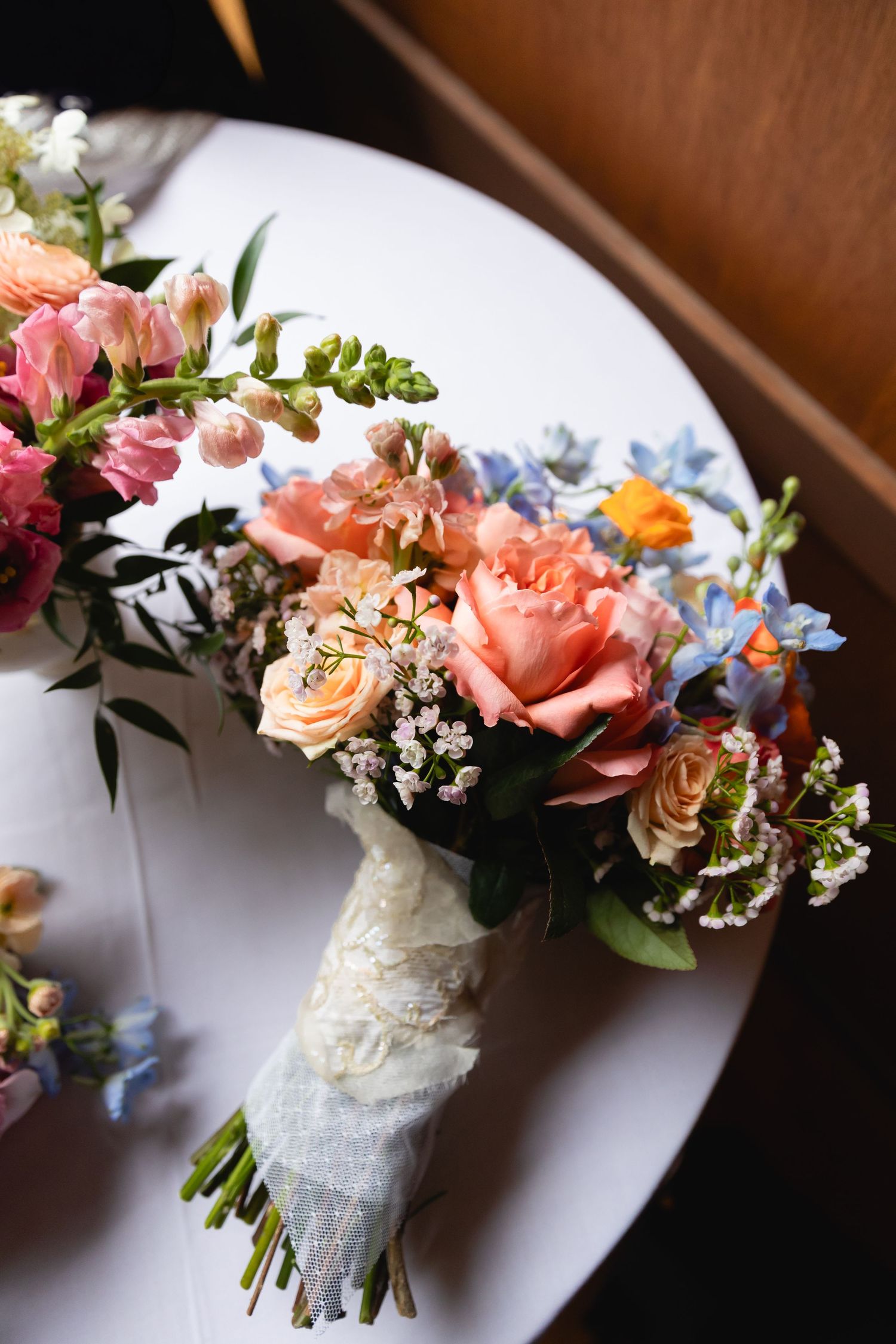 A bridal bouquet with coral roses, pink flowers, and blue delphinium wrapped in lace ribbon rests on a white plate.