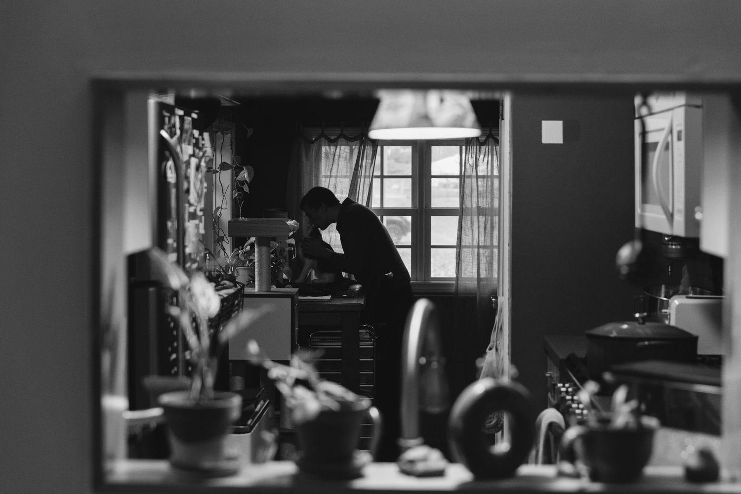 A black and white photograph showing a figure working at a desk by a window in a dimly lit interior.