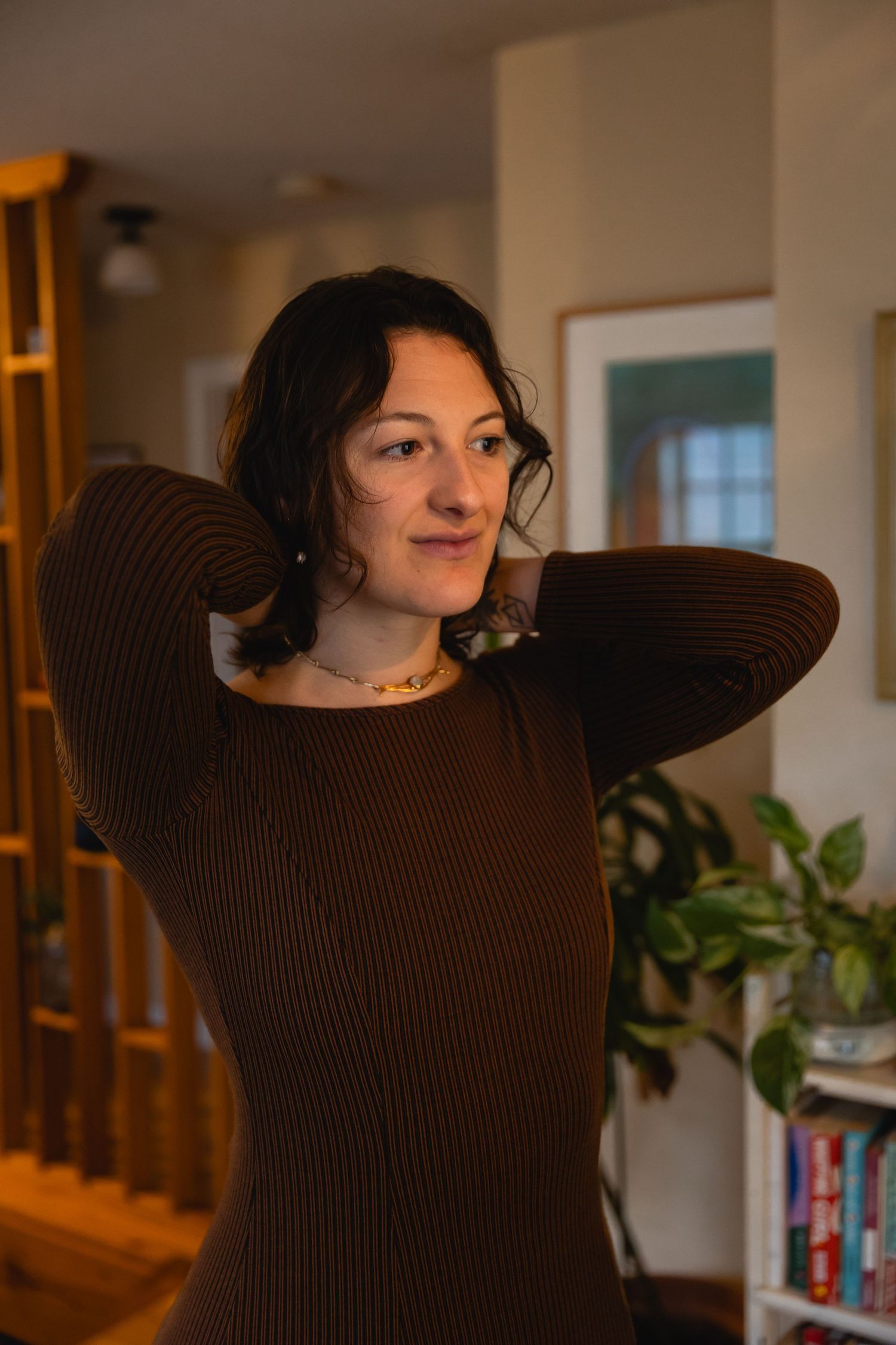 Person in a brown ribbed sweater poses casually in a cozy home setting with warm lighting and houseplants.