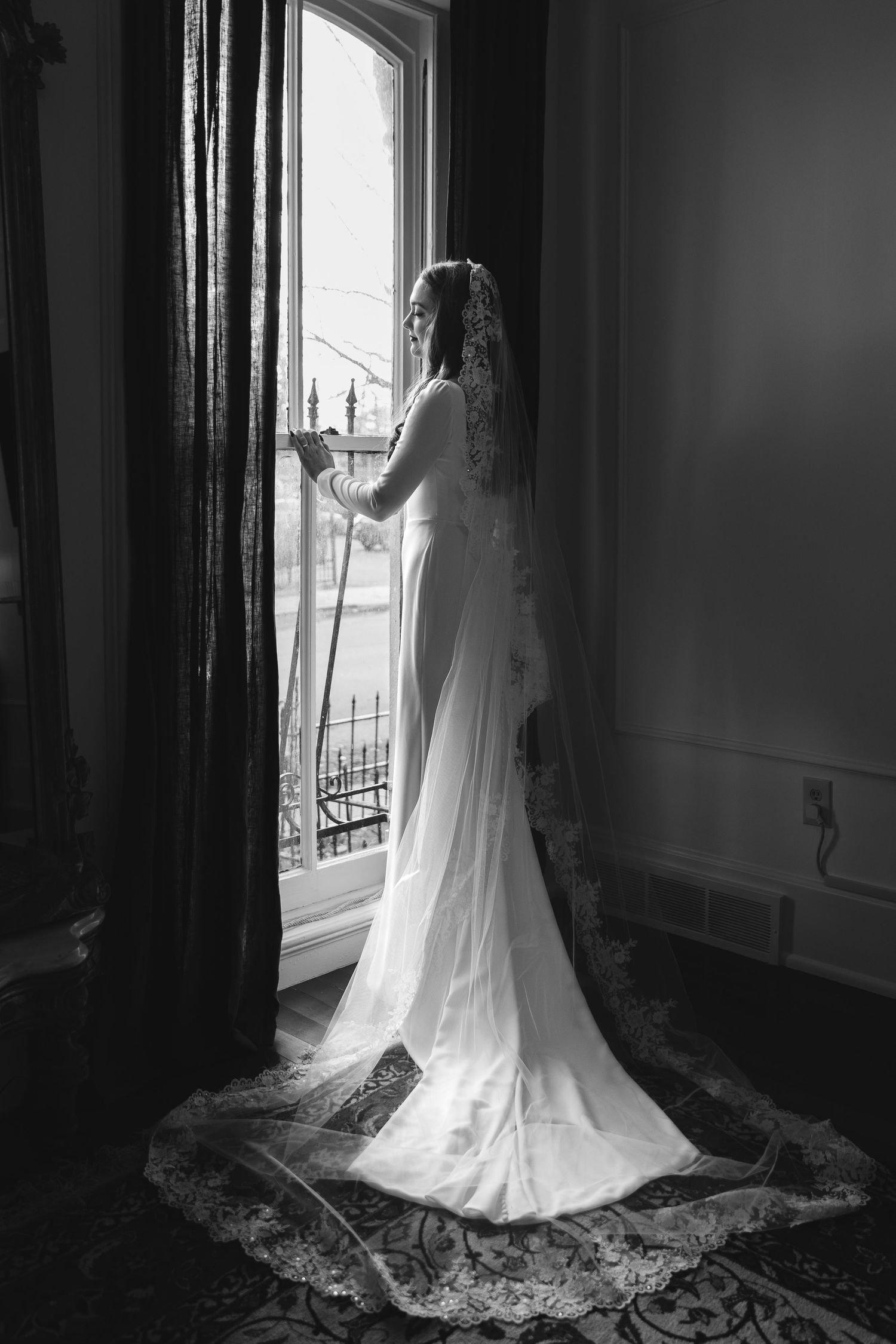 Dramatic black and white silhouette of a bride in full-length gown and veil standing by a vintage window.