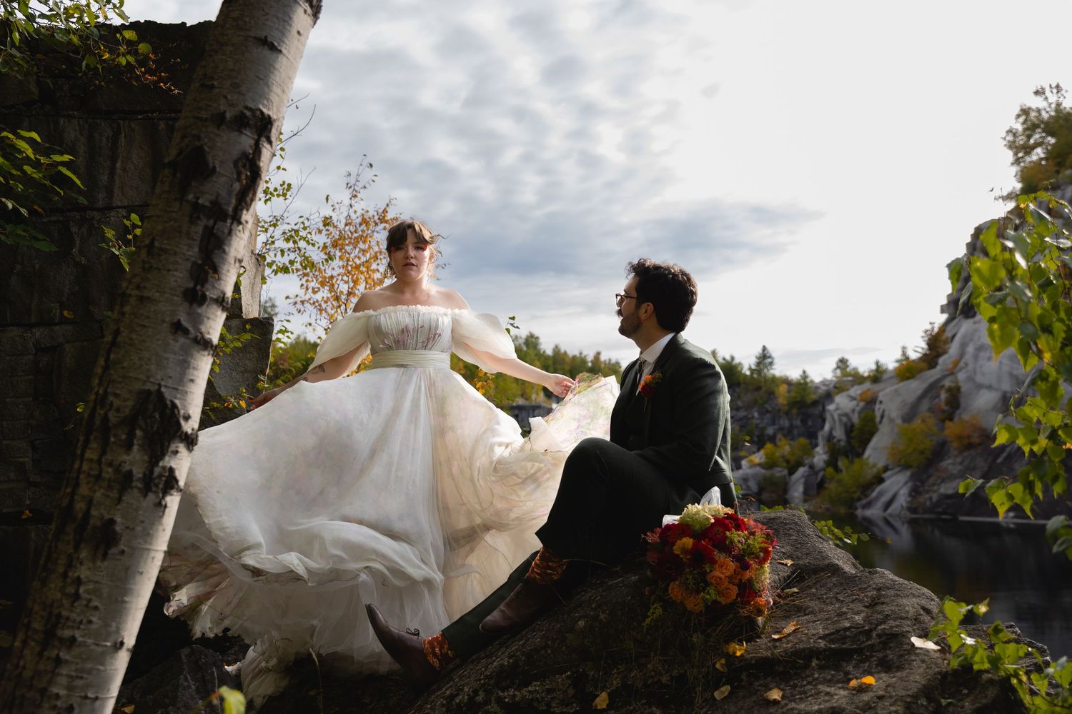 Couple in wedding attire sits on rocky ledge near birch trees during autumn with dramatic cloudy sky.