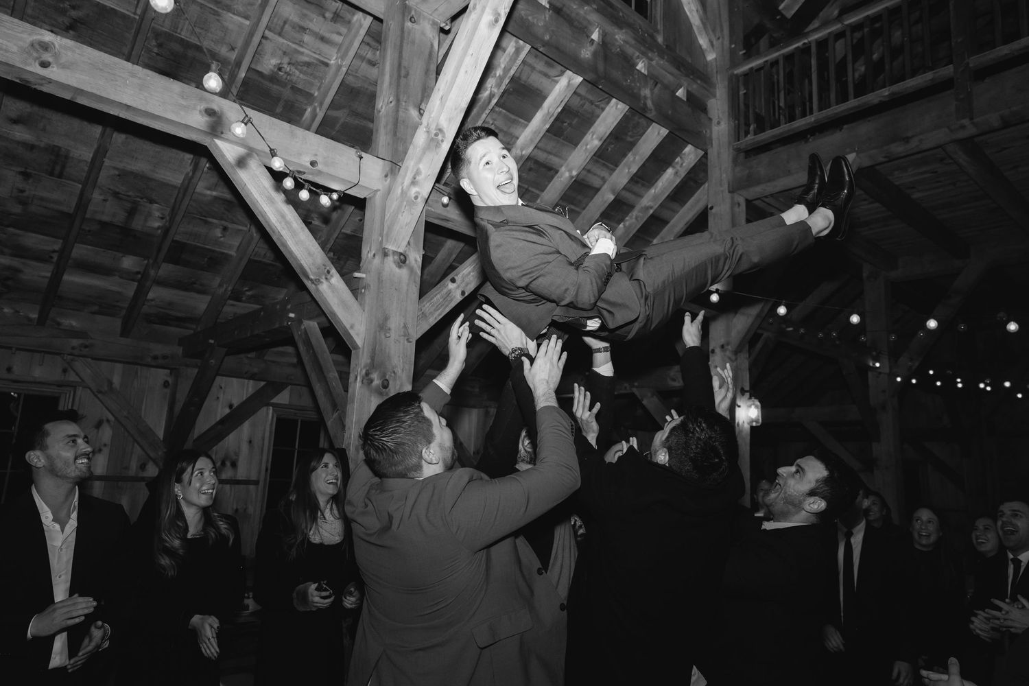 People celebrating and dancing at a rustic barn venue with exposed wooden beams and string lights overhead.