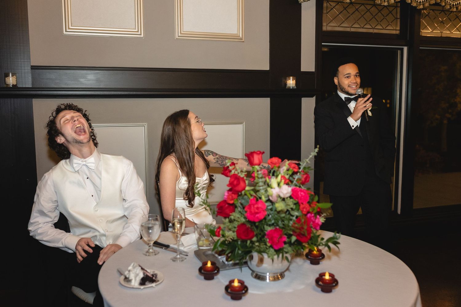 Wedding guests enjoy candid moment at reception table with centerpiece of red and pink roses during speech.