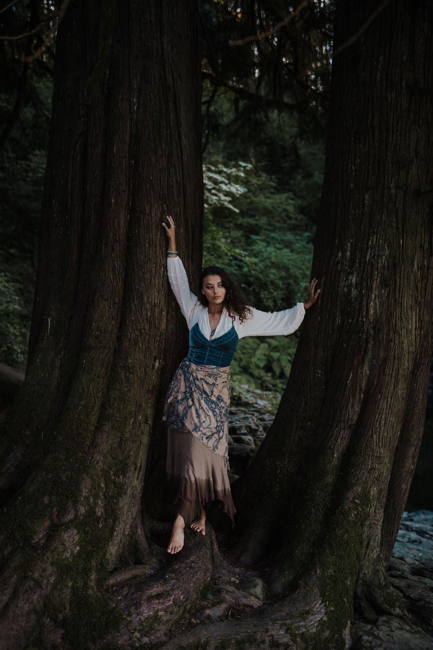 Barefoot woman in vintage attire standing between massive tree trunks in lush forest.