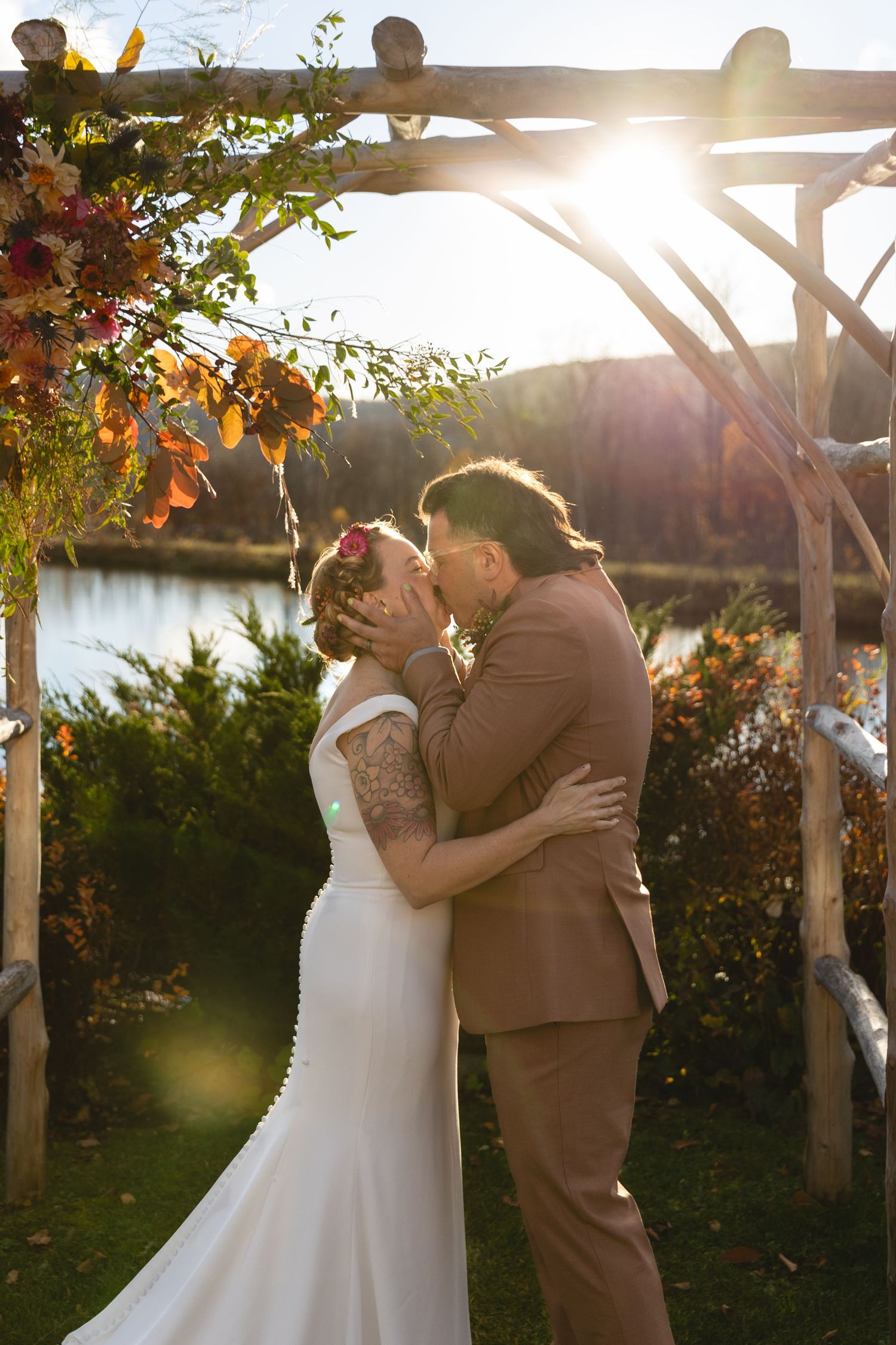 Bride and groom share first kiss under a rustic wooden arch adorned with fall foliage by a sunlit lake.