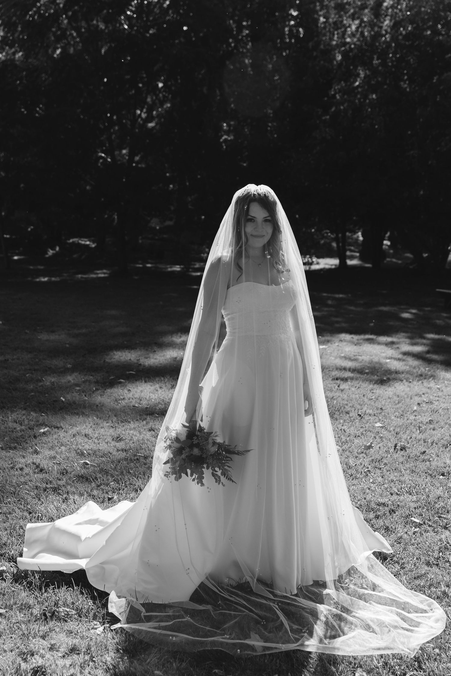 Black and white portrait of bride in white gown and veil holding bouquet outdoors.