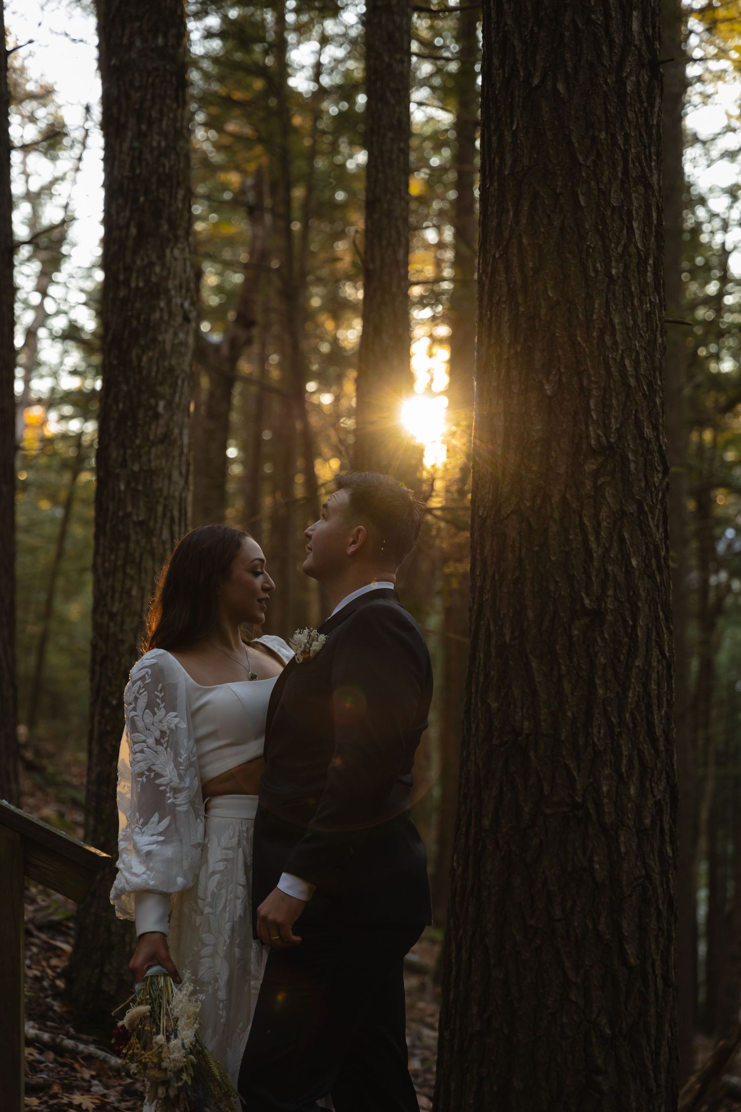Bride and groom standing among tall trees with golden sunset light streaming through the forest.