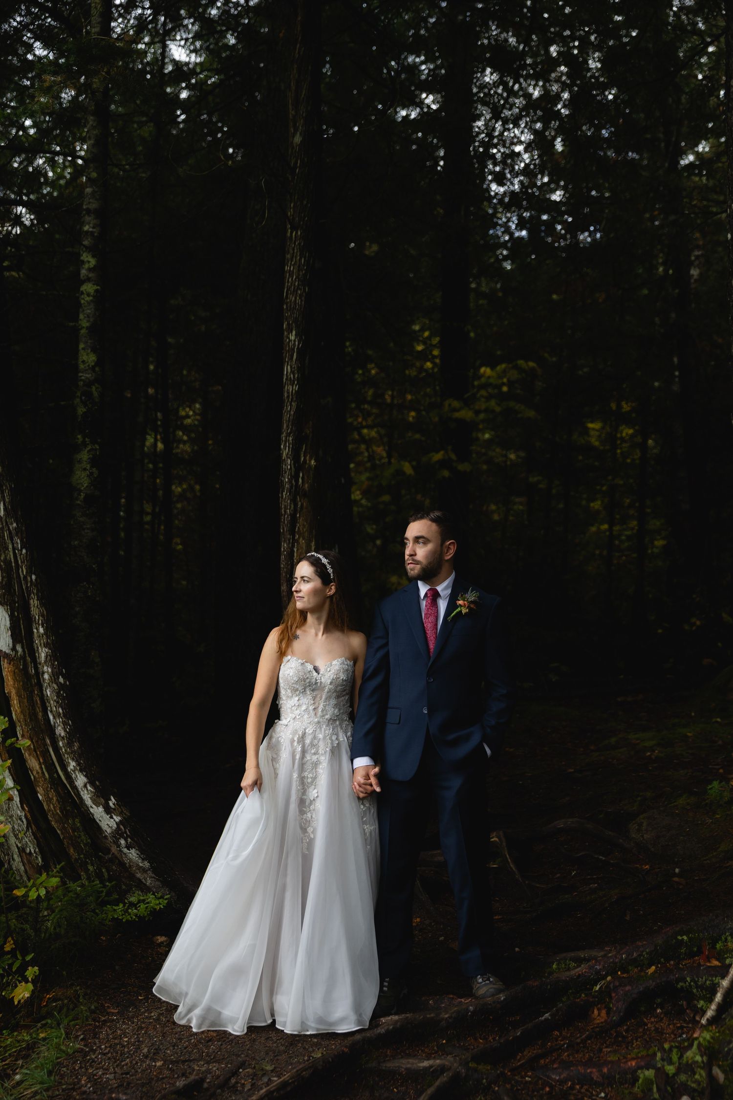 Bride in lace gown and groom in navy suit hold hands in a dark forest setting.