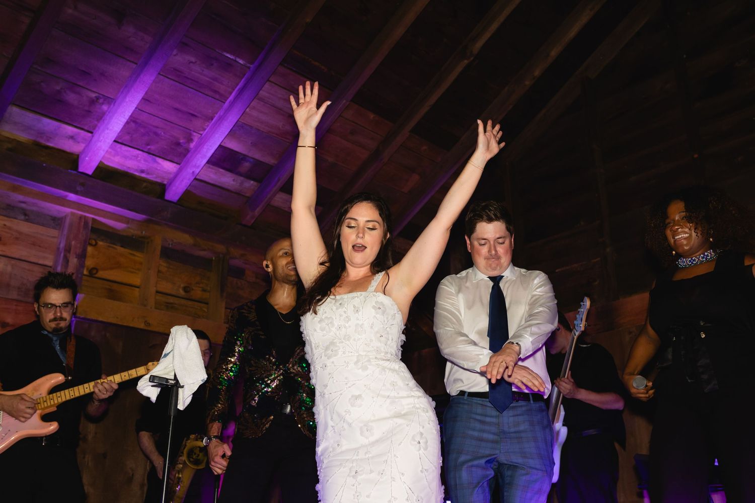 Wedding reception dancers raise their hands with joy as band performs under purple lighting at rustic barn venue.