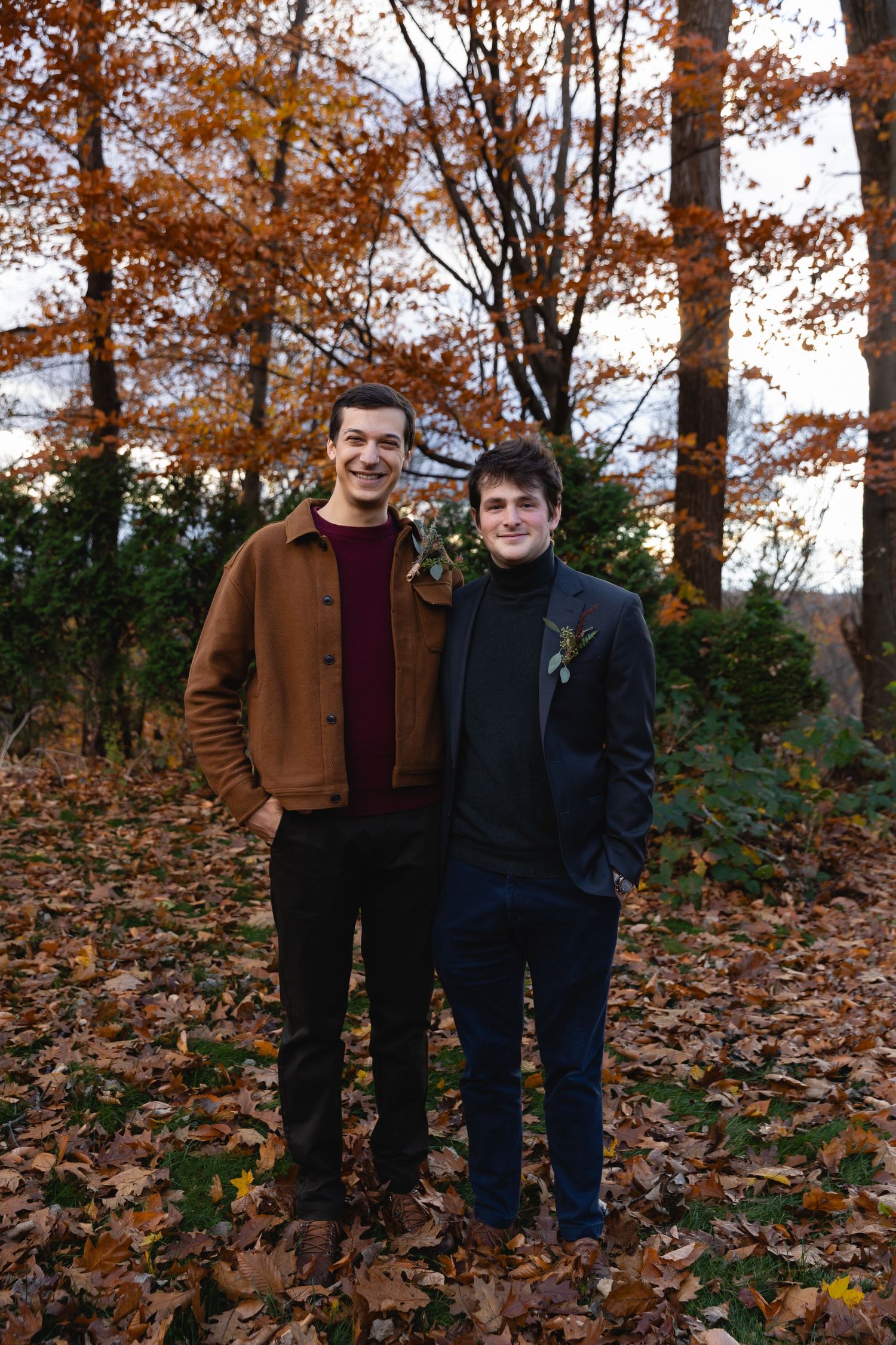 Two people standing together on fallen autumn leaves in a forest with orange and brown foliage in the background.