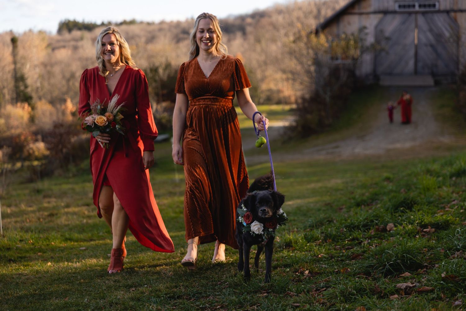 Two people in flowing red dresses walk a black dog through an autumn field at sunset with a barn in background.