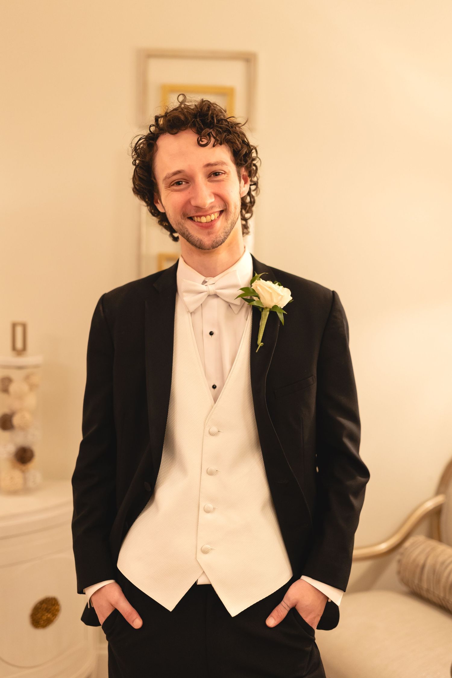 A groom wearing a black tuxedo with white vest and bowtie smiles warmly while standing with hands in pockets.