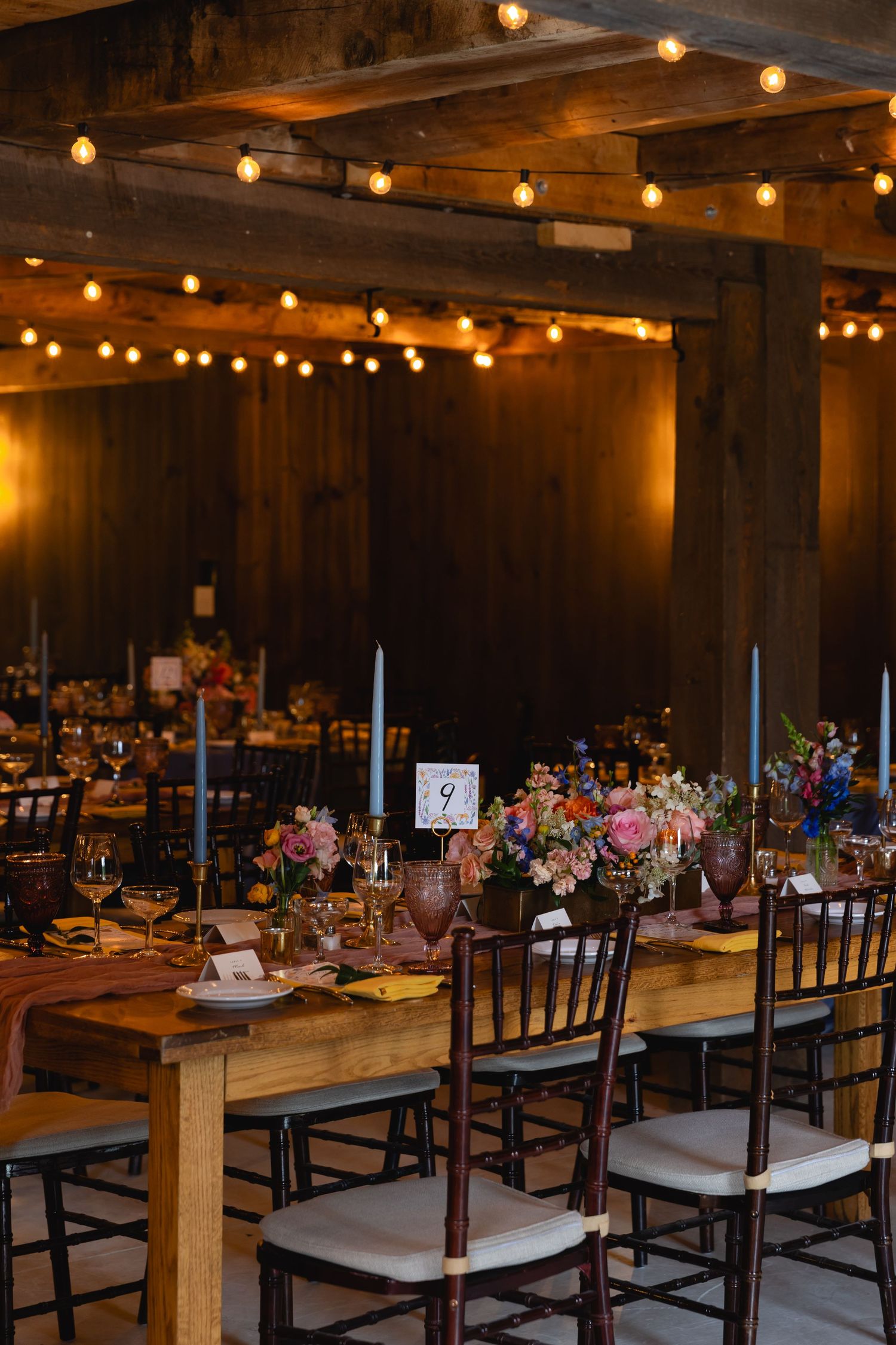 Rustic wedding reception venue with wooden tables, Chiavari chairs, string lights and colorful floral centerpieces.
