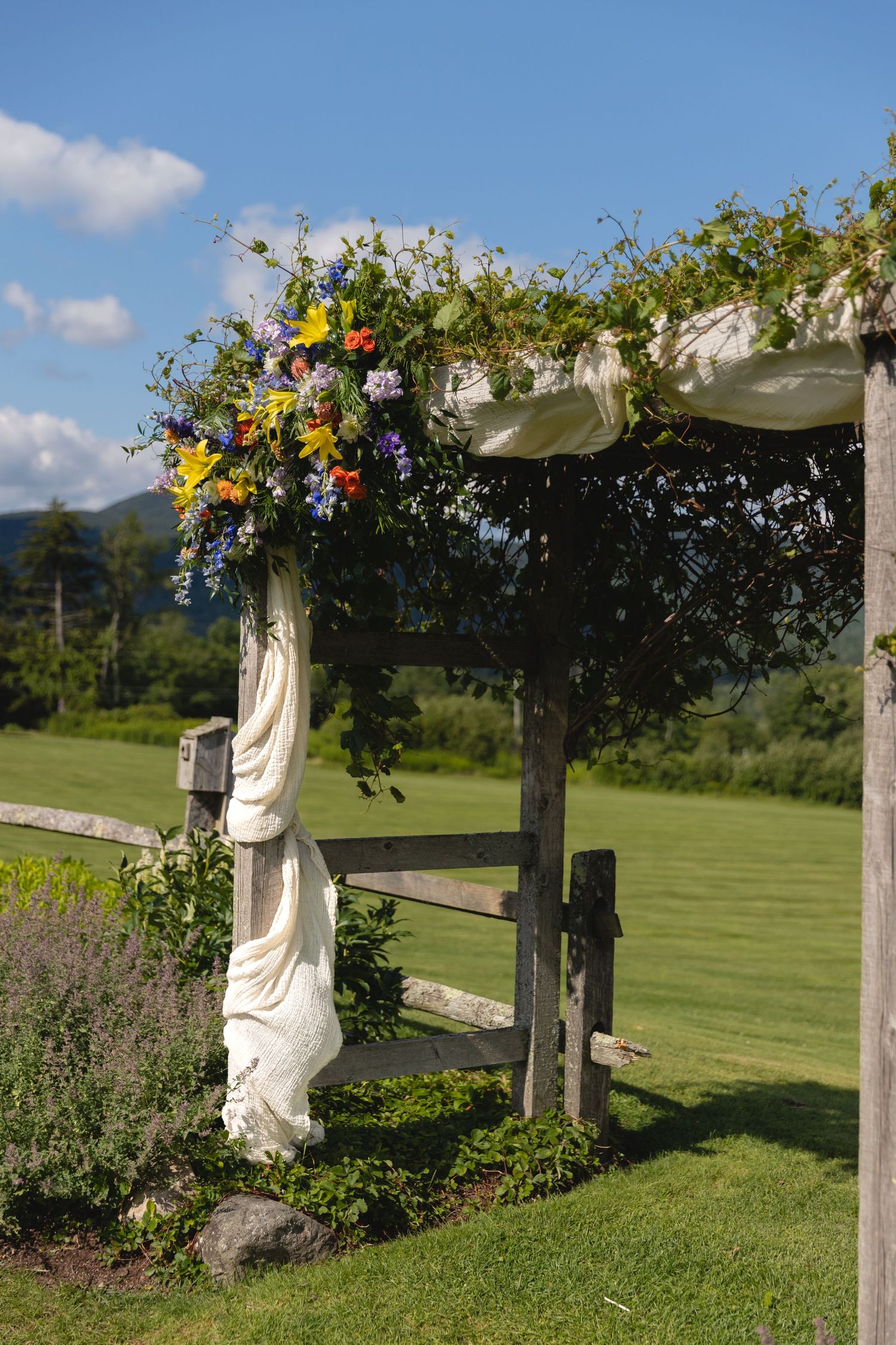 Wedding arch decorated with colorful wildflowers against mountain landscape and blue sky.