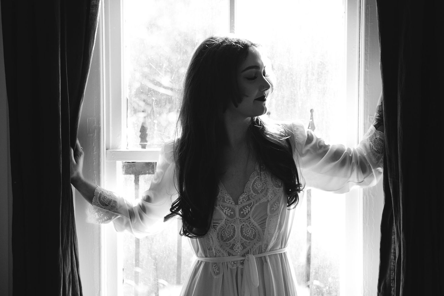 A silhouetted figure in a white lace dress stands at a bright window in a black and white photograph.
