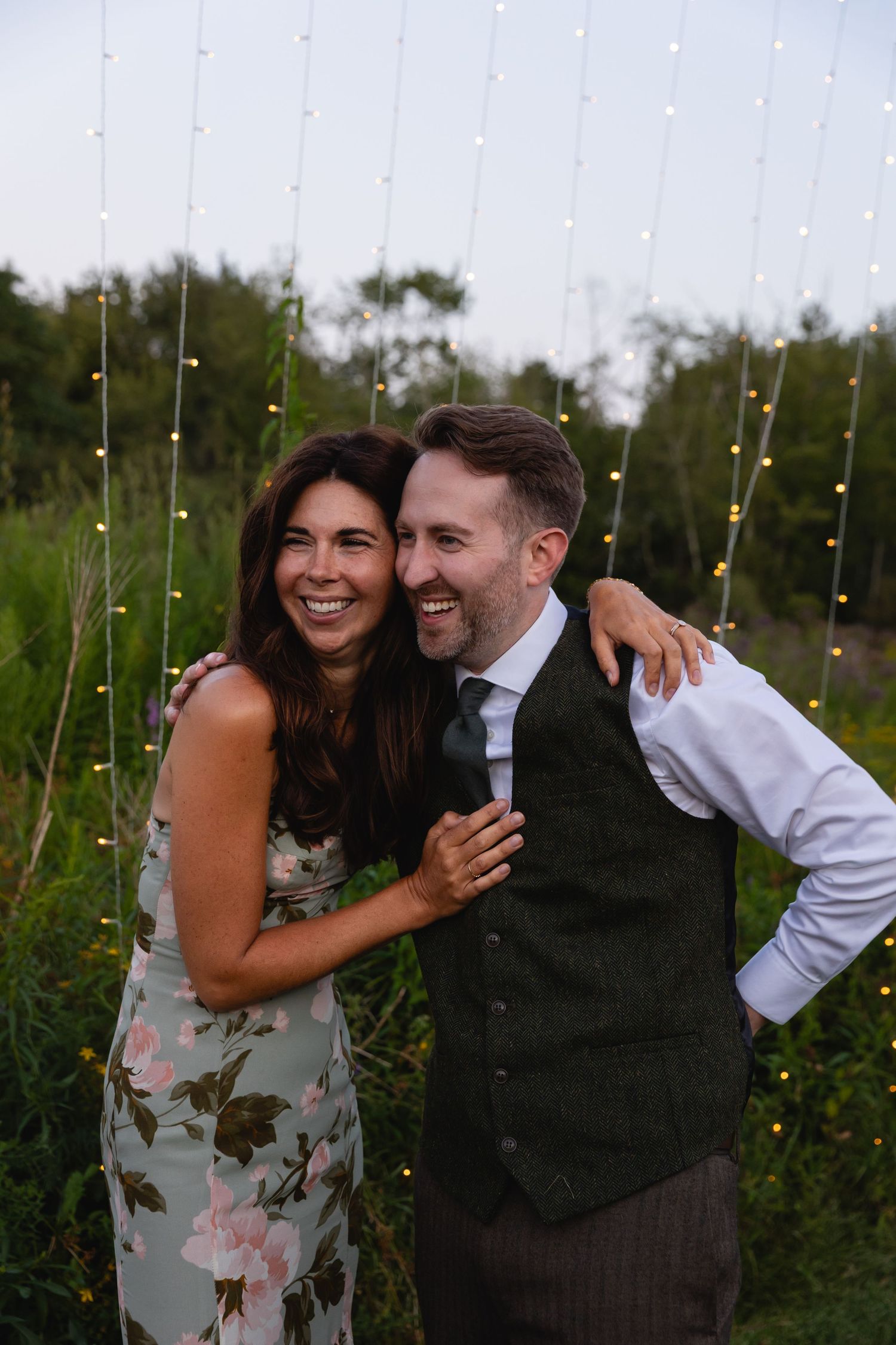 A couple shares joyful laughter together outdoors with string lights and greenery in the background.