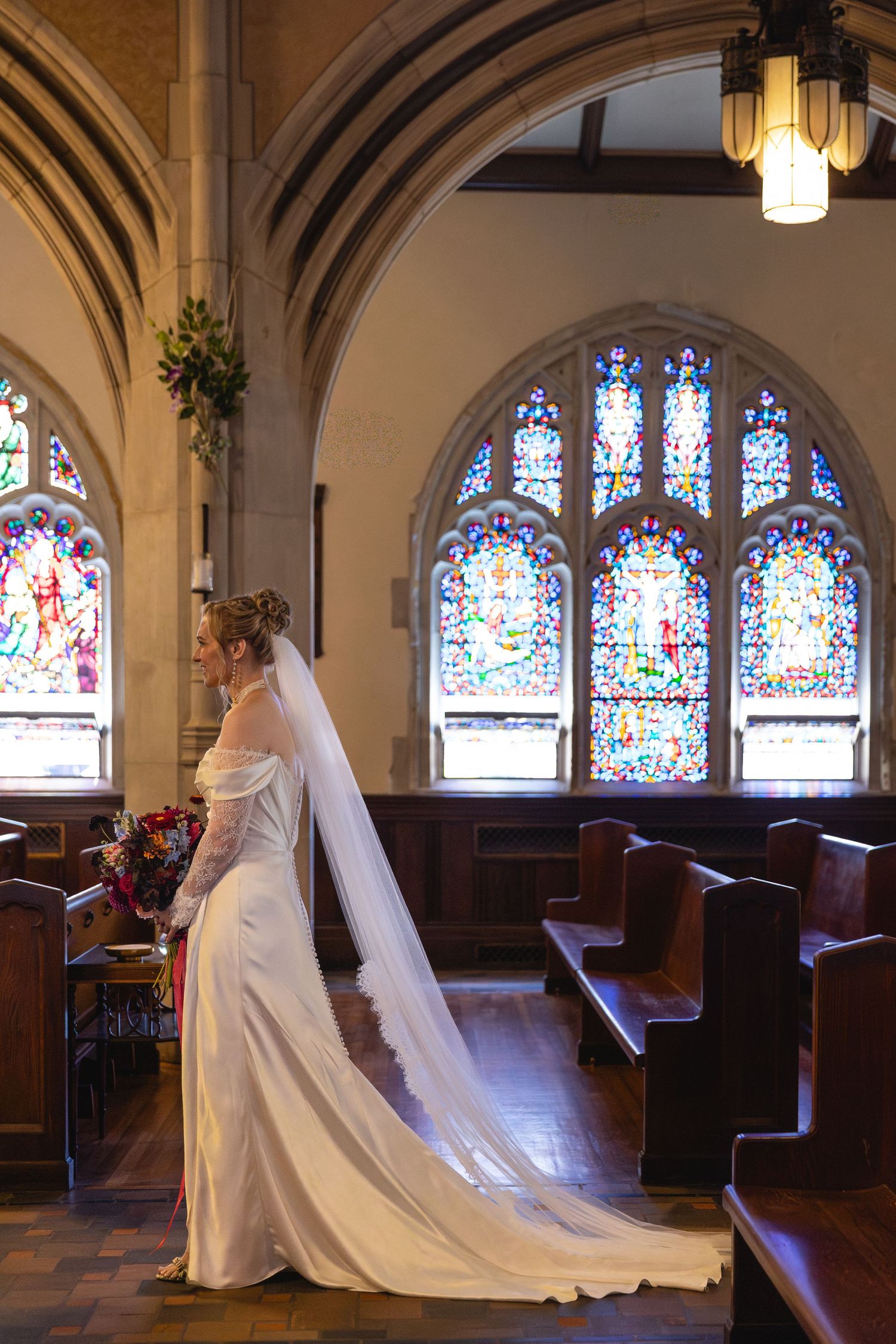 A bride in flowing white gown and veil poses in a church sanctuary with stained glass windows and wooden pews.