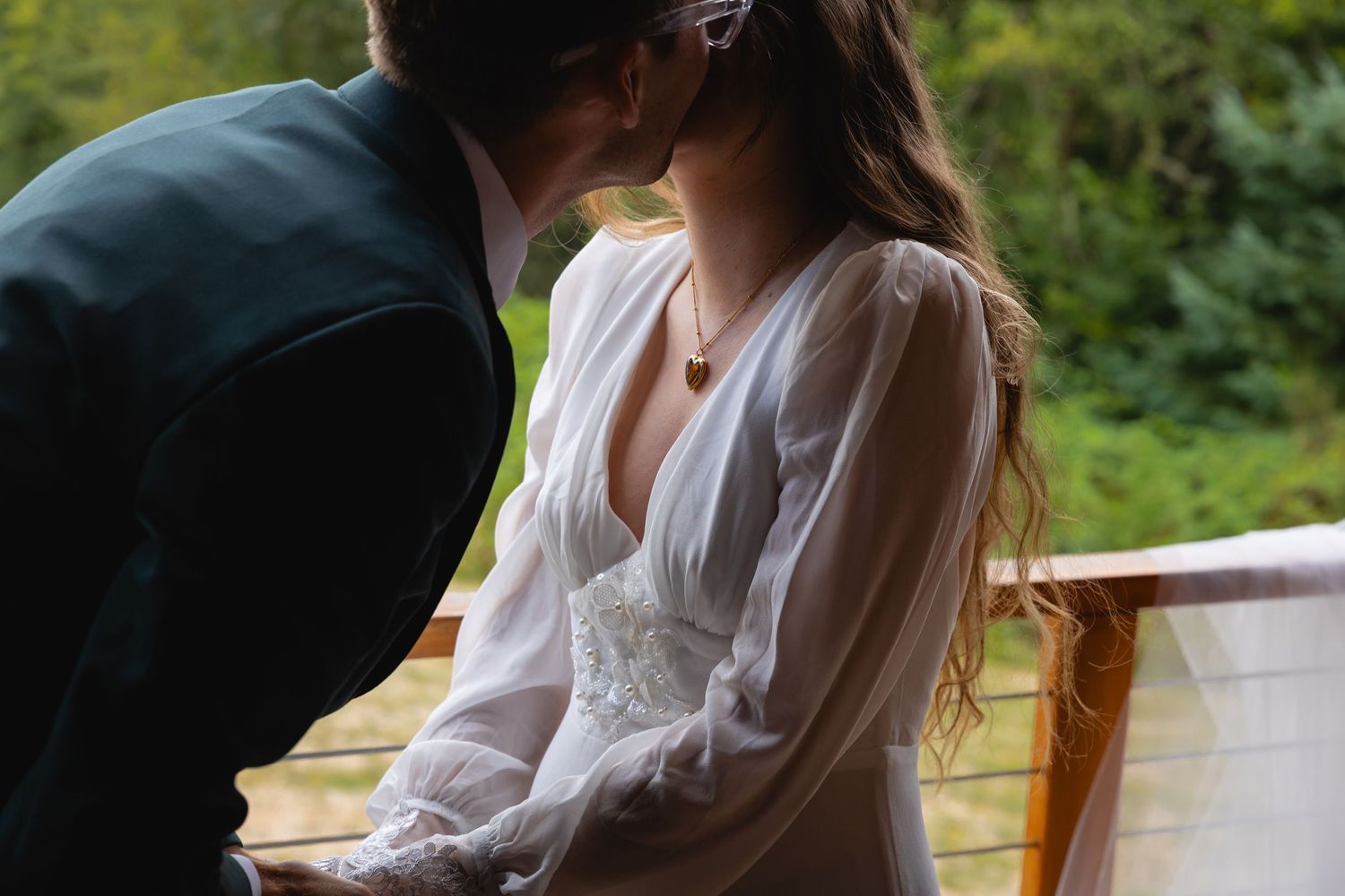 A couple shares a tender moment outdoors with sunlight filtering through trees in the background.