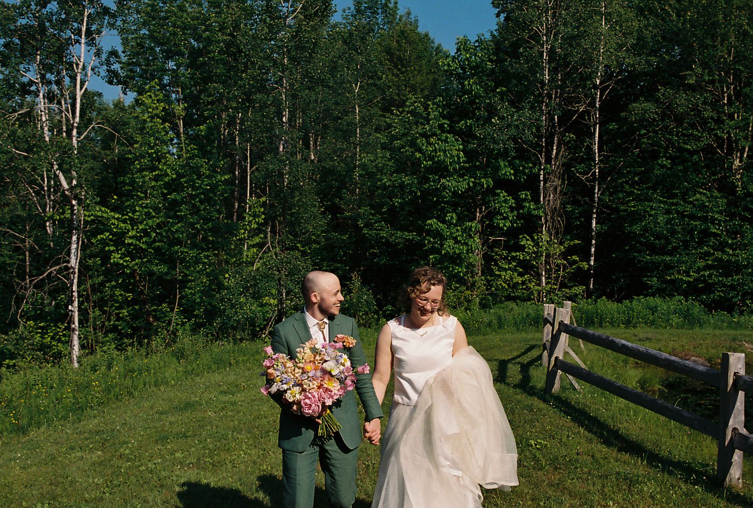 A couple walks through a lush green meadow on a sunny summer day with tall trees in the background.