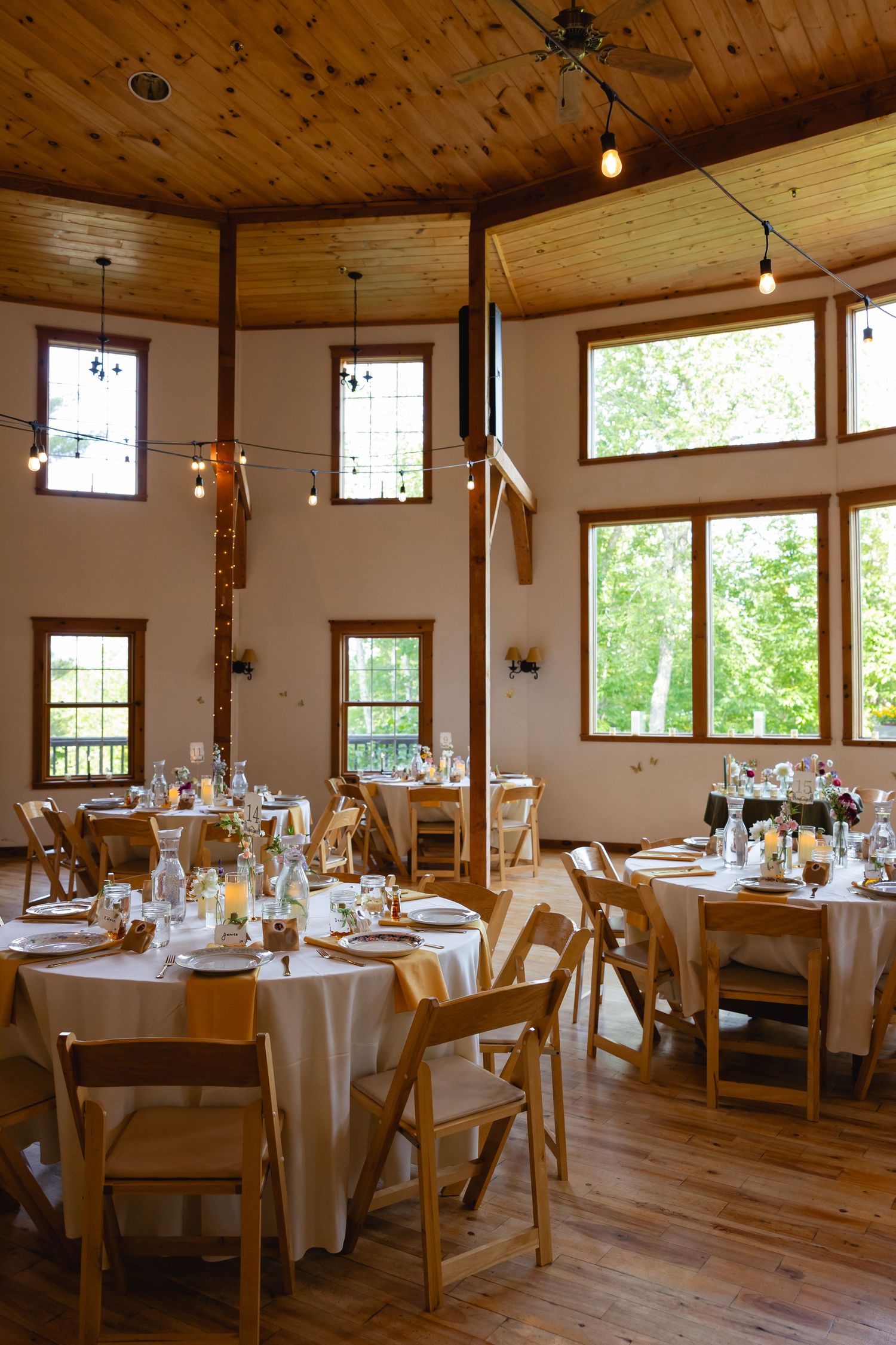 Rustic event venue interior with wooden ceiling, round tables, and natural lighting through large windows.