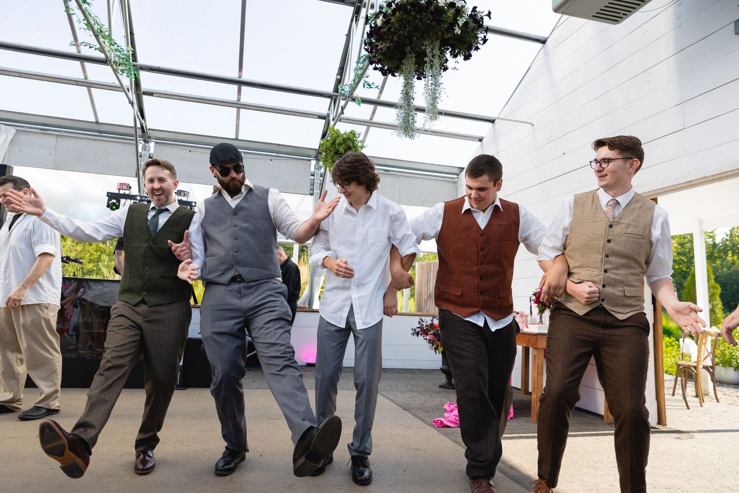 Groomsmen in coordinating vests dance together at an indoor-outdoor wedding venue under a glass ceiling.
