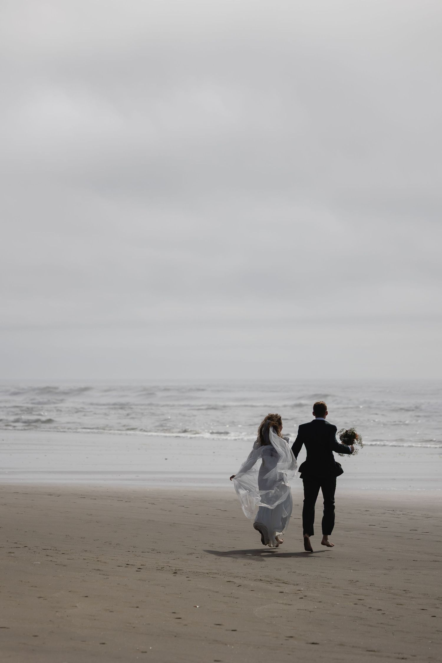 A couple in formal wedding attire walks together along a misty beach shoreline in black and white tones.