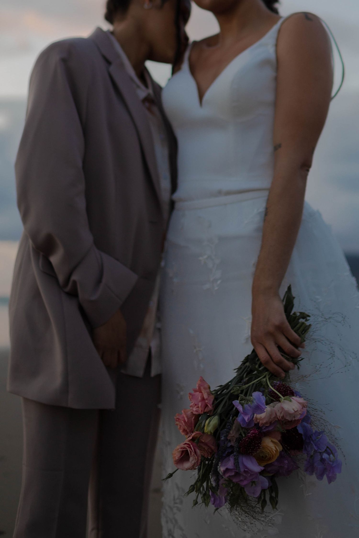 A close-up wedding photo showing elegant attire and a colorful bouquet of purple and pink flowers.