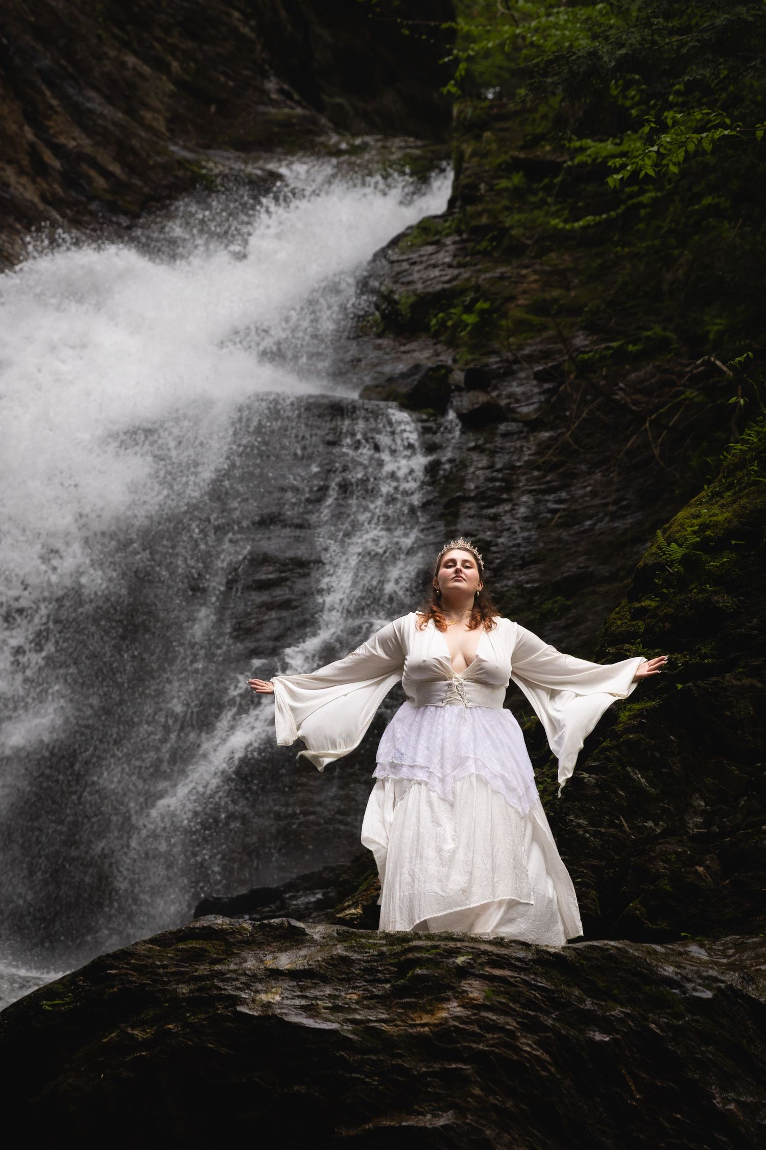 A woman in a white medieval gown stands with arms outstretched before a powerful waterfall.
