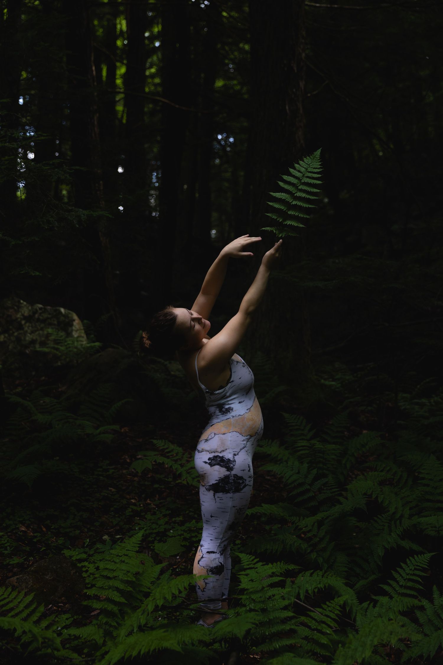 Woman in patterned athletic wear holds a fern leaf overhead in a dark moody forest setting.