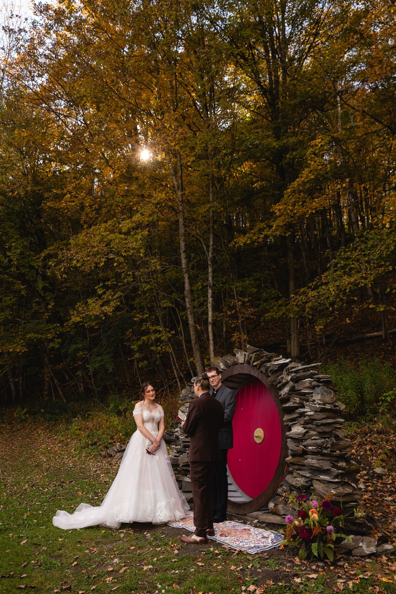 A bride and groom exchange vows before a hobbit-style stone door arch amid vibrant fall foliage.