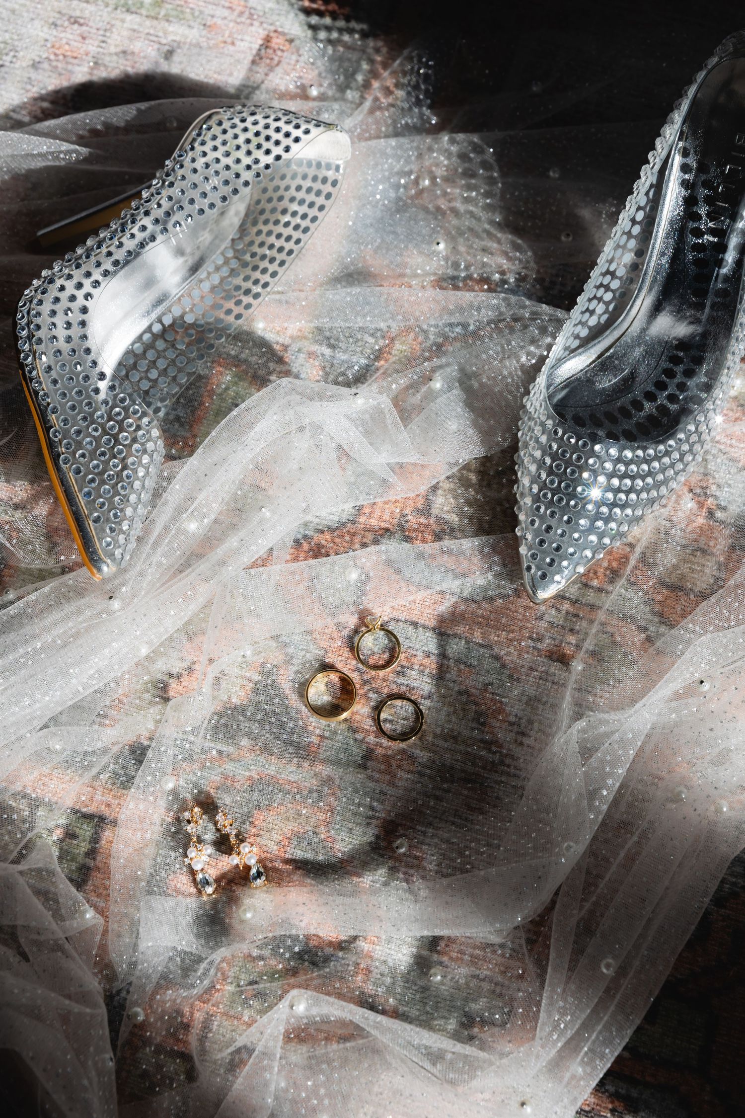 Sparkling silver high heel shoes with wedding rings and veil on dark textured surface.