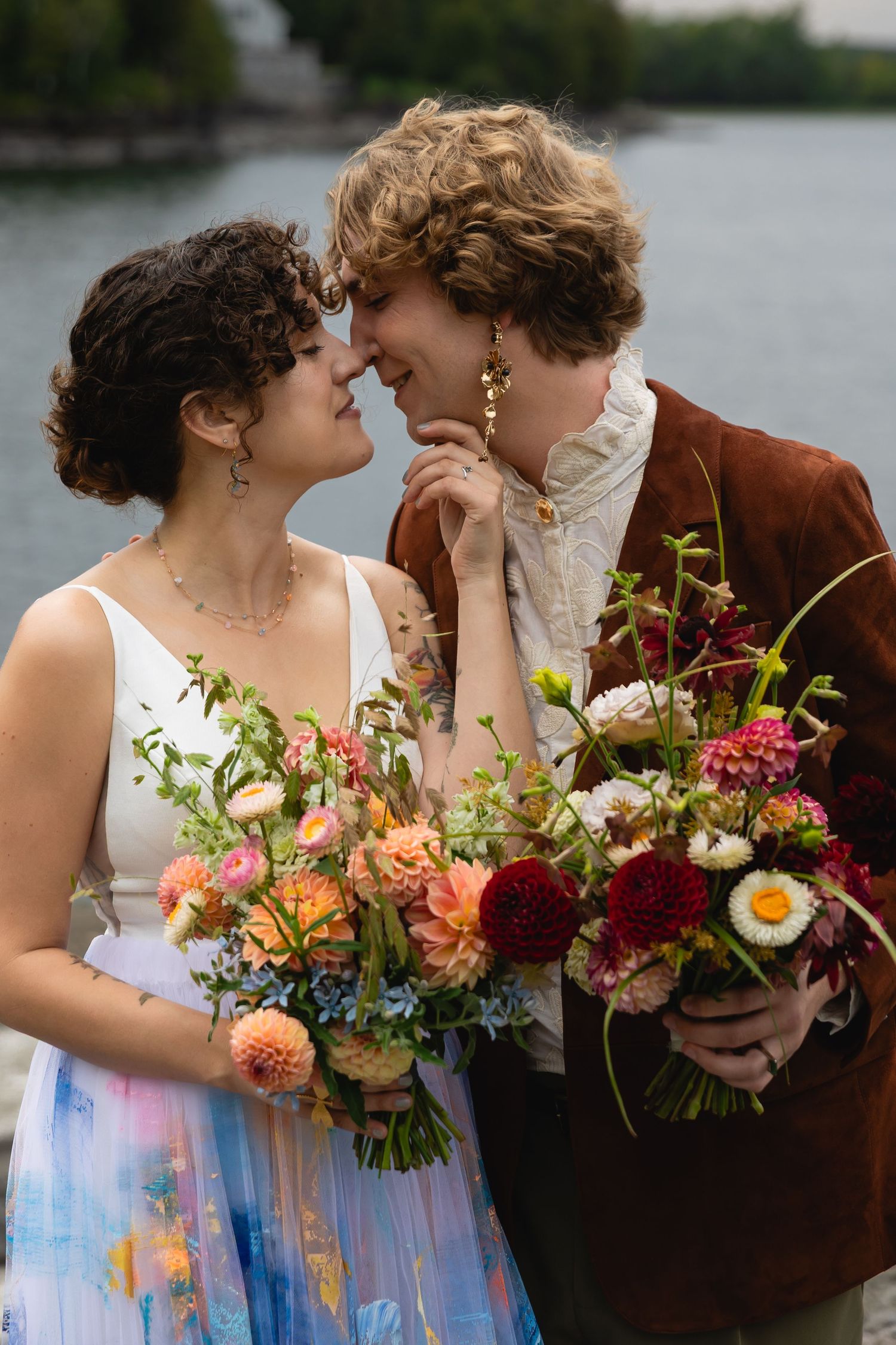 Couple nearly kissing while holding colorful wildflower bouquets near a waterfront on their wedding day.