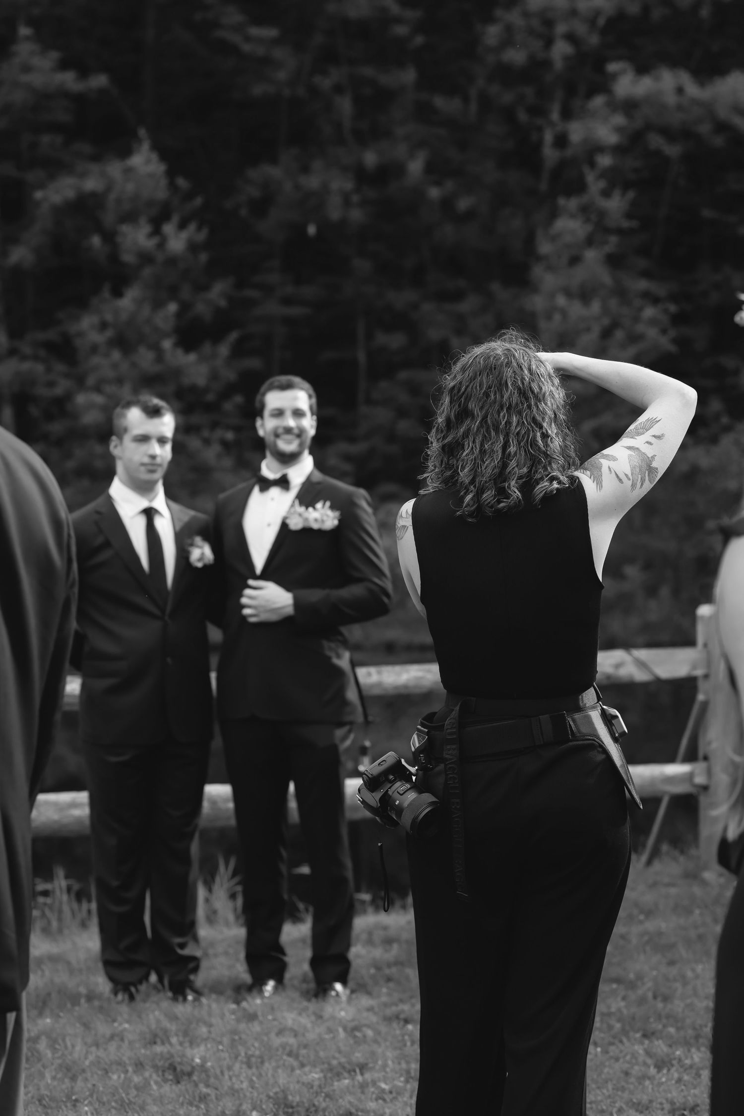 Black and white photo of female photographer directing smiling groom and groomsman at wedding.